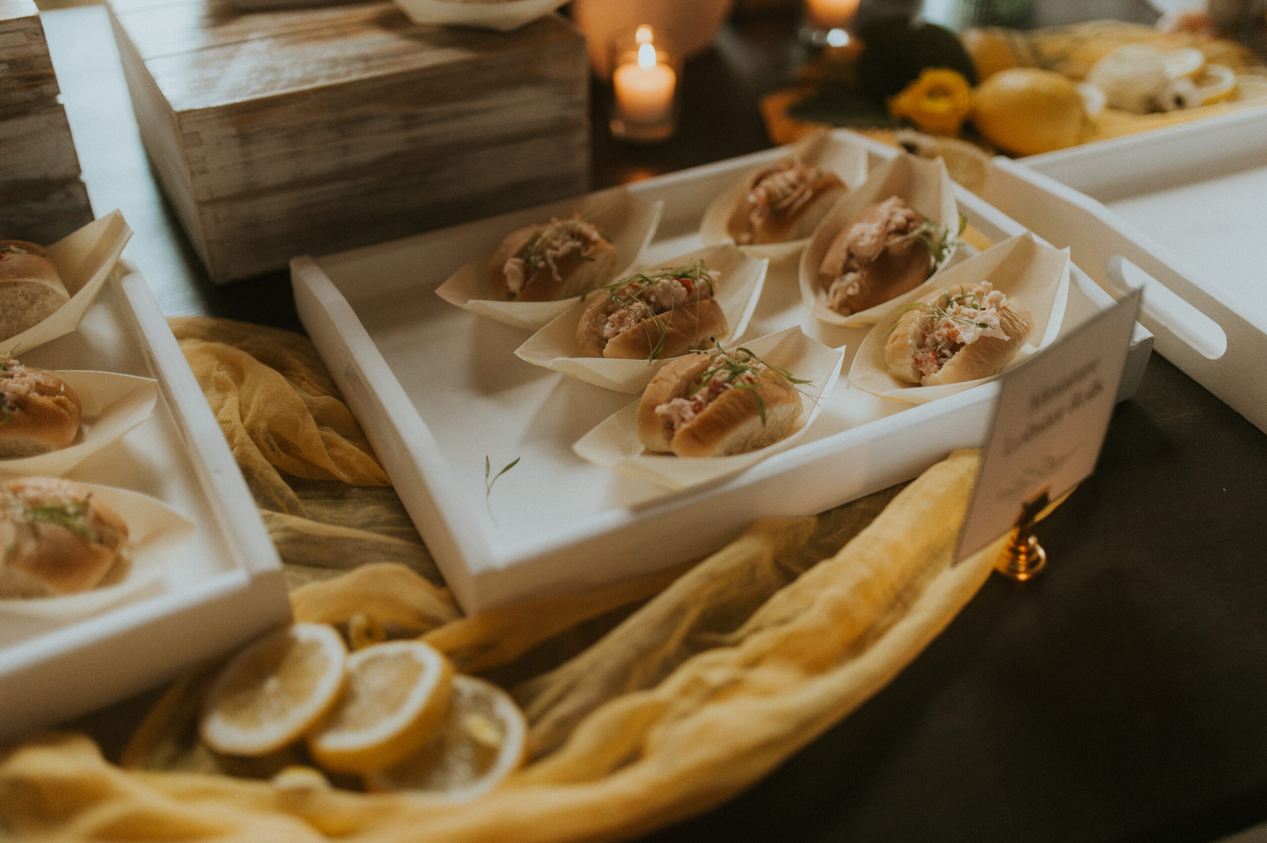 Miniature Lobster Roll Appetizer Stationary display of miniature lobster rolls beautifully arranged on a table with a yellow runner and fresh lemons, creating an elegant and vibrant appetizer station for weddings and events.