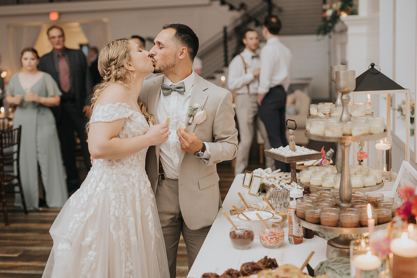 Bride and Groom Kissing in Front of Mason Jar Dessert Bar – Wedding Dessert Table Bride and groom share a kiss in front of a beautifully decorated dessert table, featuring a mason jar dessert bar with mini cheesecakes and a variety of toppings, including sprinkles, caramel syrup, and chocolate syrup.
