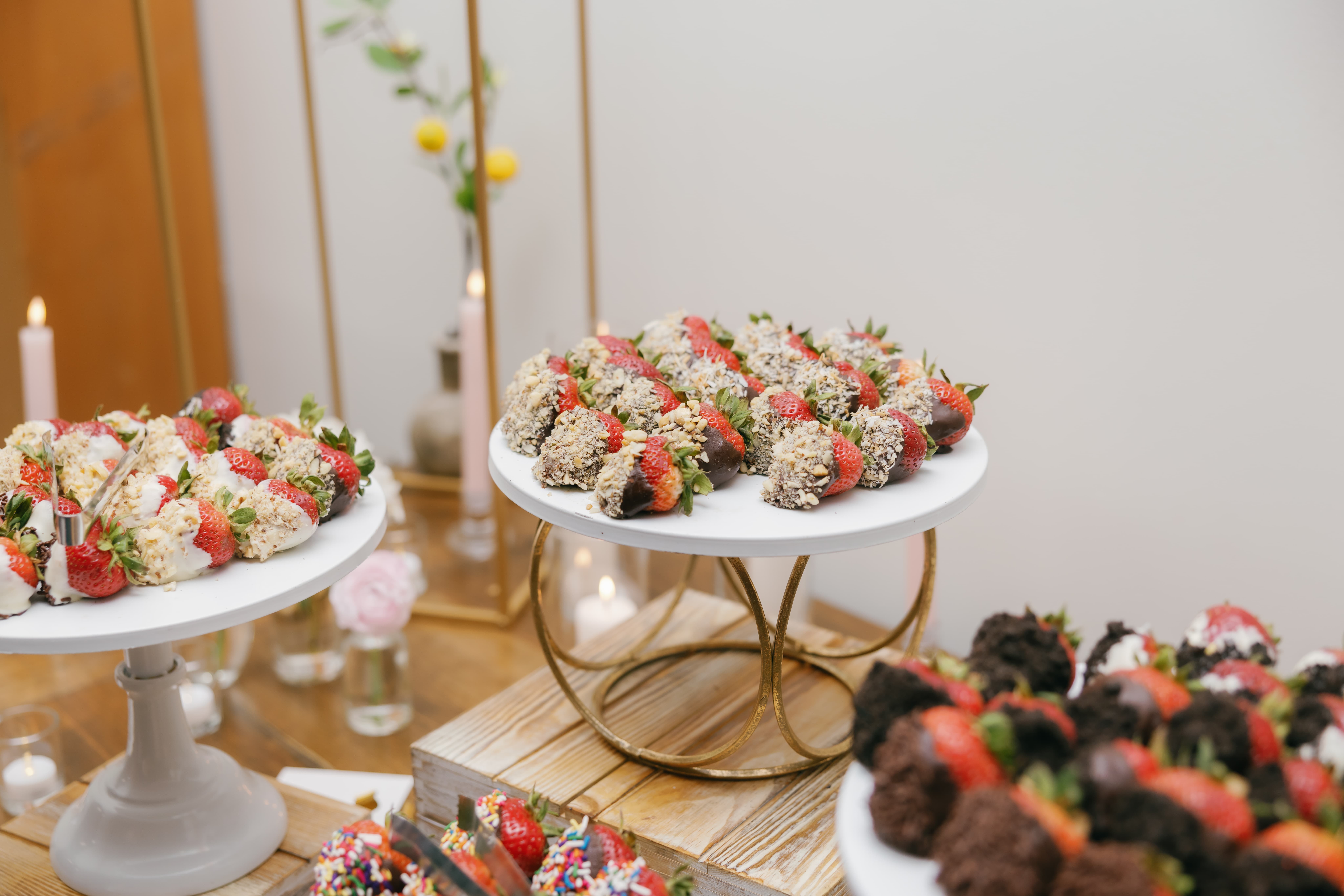 Chocolate Covered Strawberries A display of fresh strawberries dipped in milk and white chocolate, coated with toasted coconut flakes, nuts, sprinkles, or crushed cookies. The dessert table is elegantly decorated with florals, pillar candles, and themed decor for a beautiful presentation.