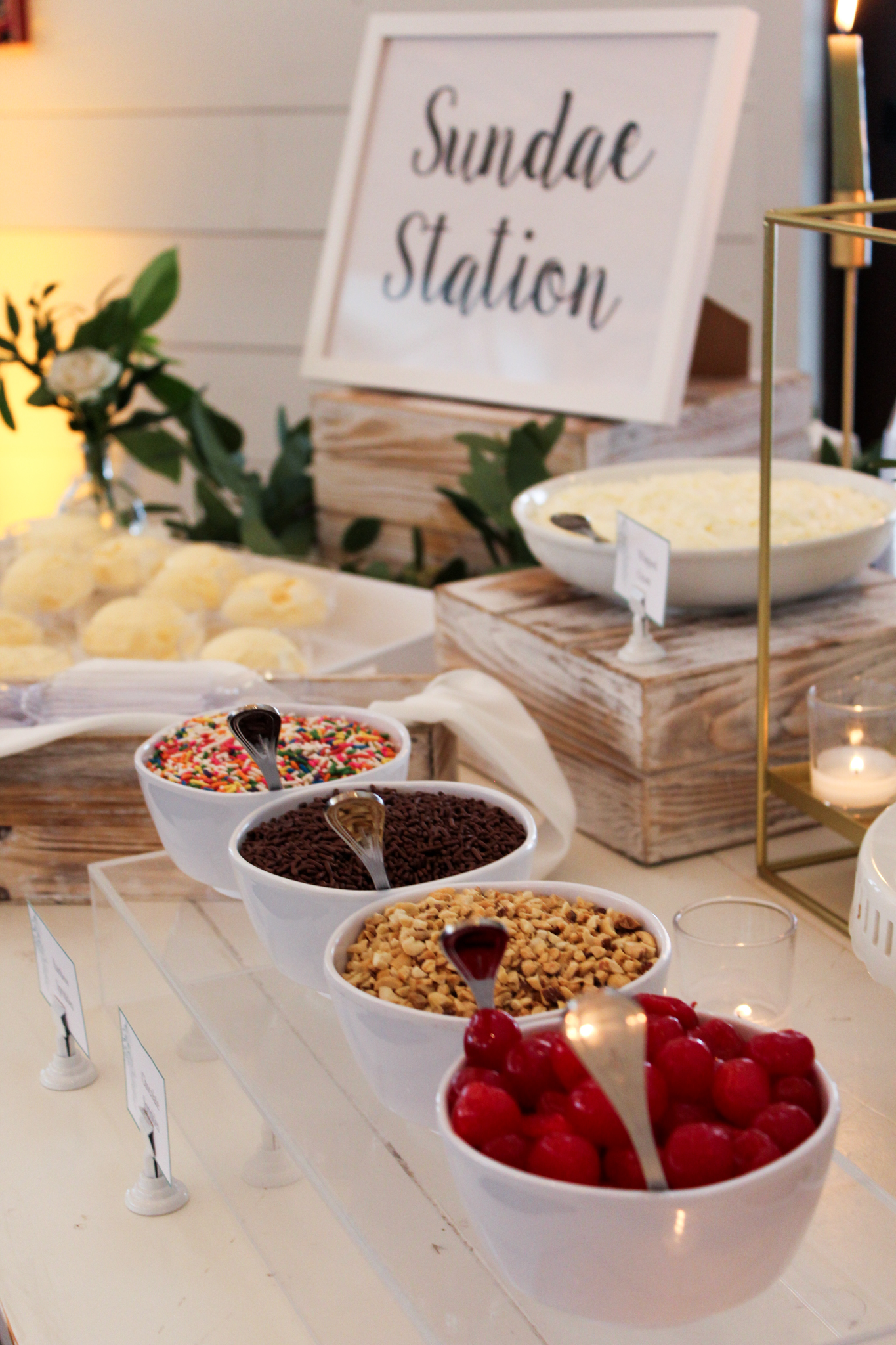 Sundae Station Sundae station featuring pre-scooped vanilla ice cream in bowls, accompanied by an assortment of toppings, including brownies, chocolate sauce, caramel sauce, chopped nuts, sprinkles, whipped cream, and cherries. The dessert table is decorated for an elegant presentation.