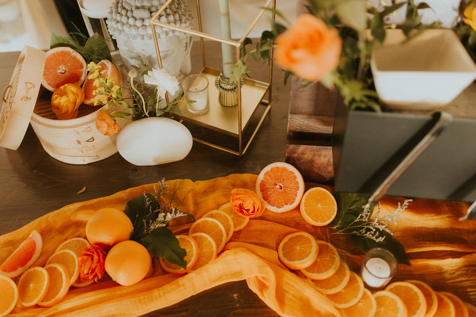 Asian Noodle Bar Station Table Decor A warm, citrus-themed table display with sliced oranges, grapefruits, vibrant flowers, candles, and a bamboo steamer basket on an orange fabric runner, creating a bright and rustic decorative arrangement for an Asian Noodle station.