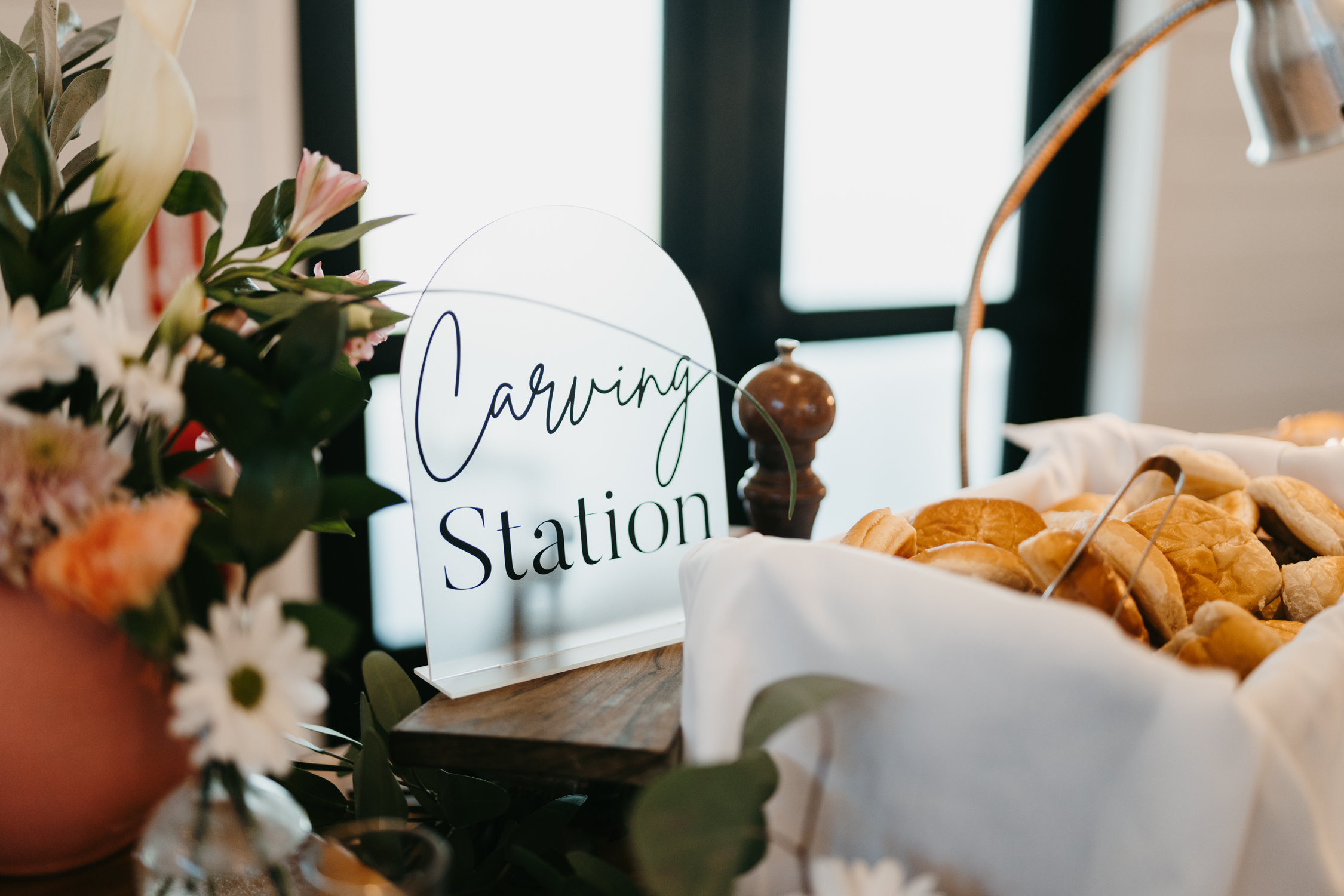 Carving Station Signage A beautifully decorated table setup for the Carving Station, including a custom acrylic sign with the name of the station. Rolls are out for the taking, and flowers cascade on the table.