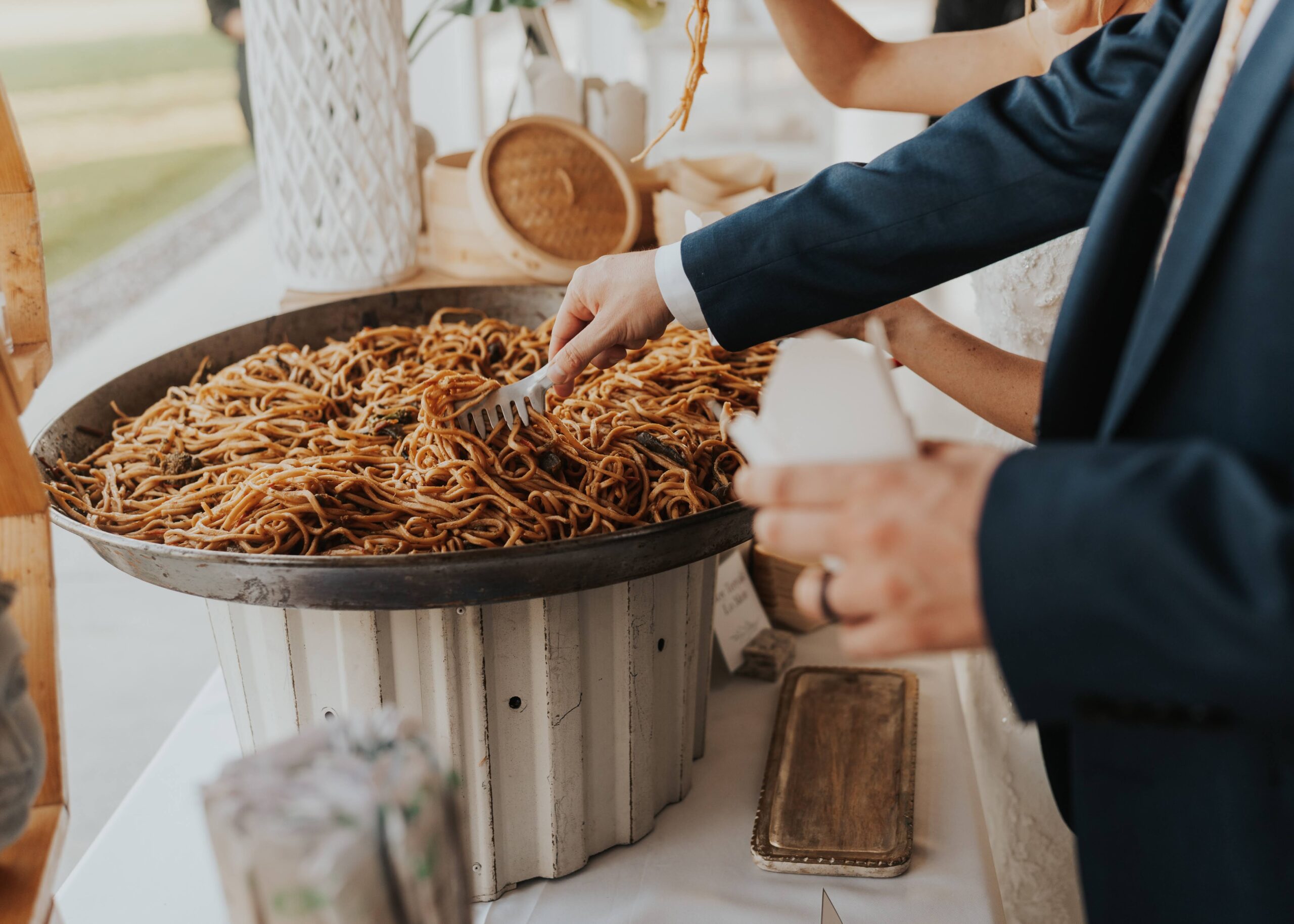 Lo Mein Noodles at Asian Noodle Station A wok full of Lo Mein Noodles atop a table for an Asian Noodle Station. The groom is reaching down and putting his food into a traditional food takeout container. The table is lined with themed decor and signage.