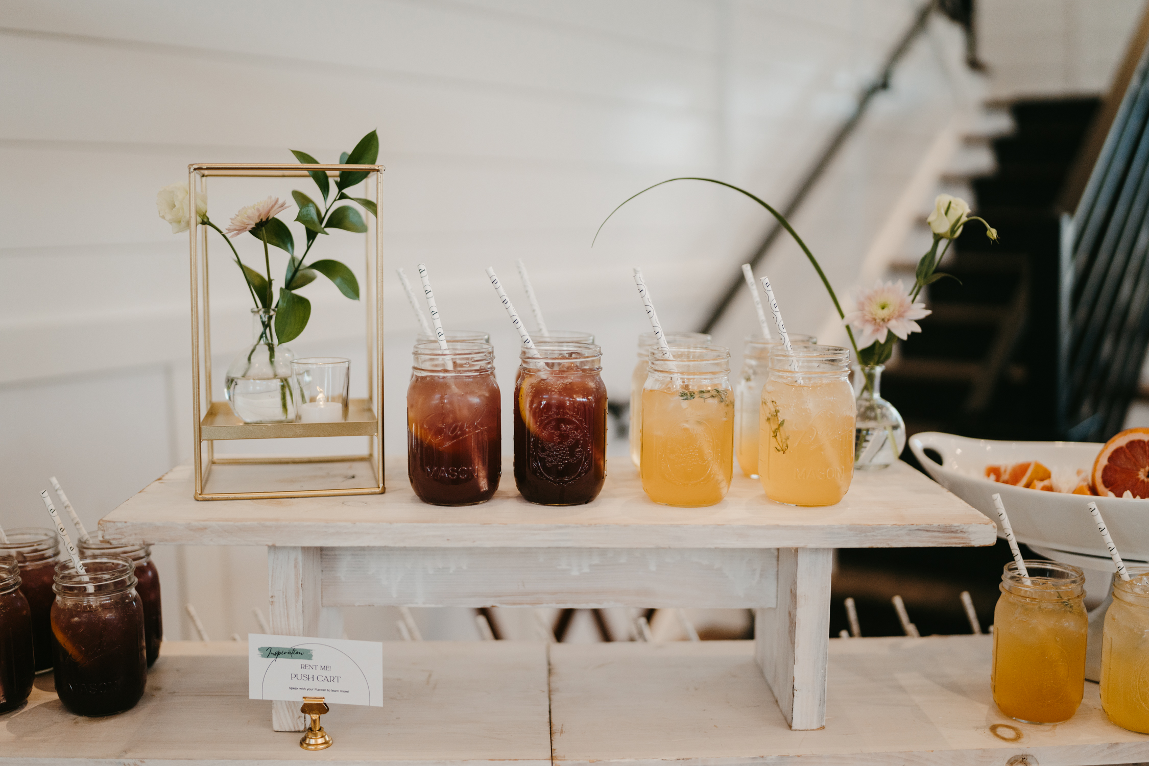 Signature Mocktails Two signature mocktails, 'Apricot Sunset Sip' and 'Blueberry Lavender Bubbles,' displayed on nesting tables with elegant decor and a fresh fruit arrangement, perfect for weddings and events.