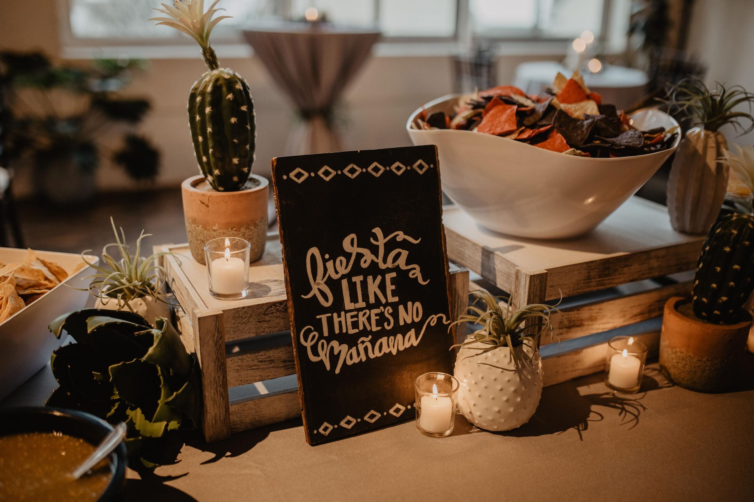 Taco Station with Signage and Decor A festive taco station setup with a sign that reads "Fiesta like there's no mañana," surrounded by cacti, votive candles, air plants, salsa, and bowls of colorful tortilla chips on rustic wooden crates.