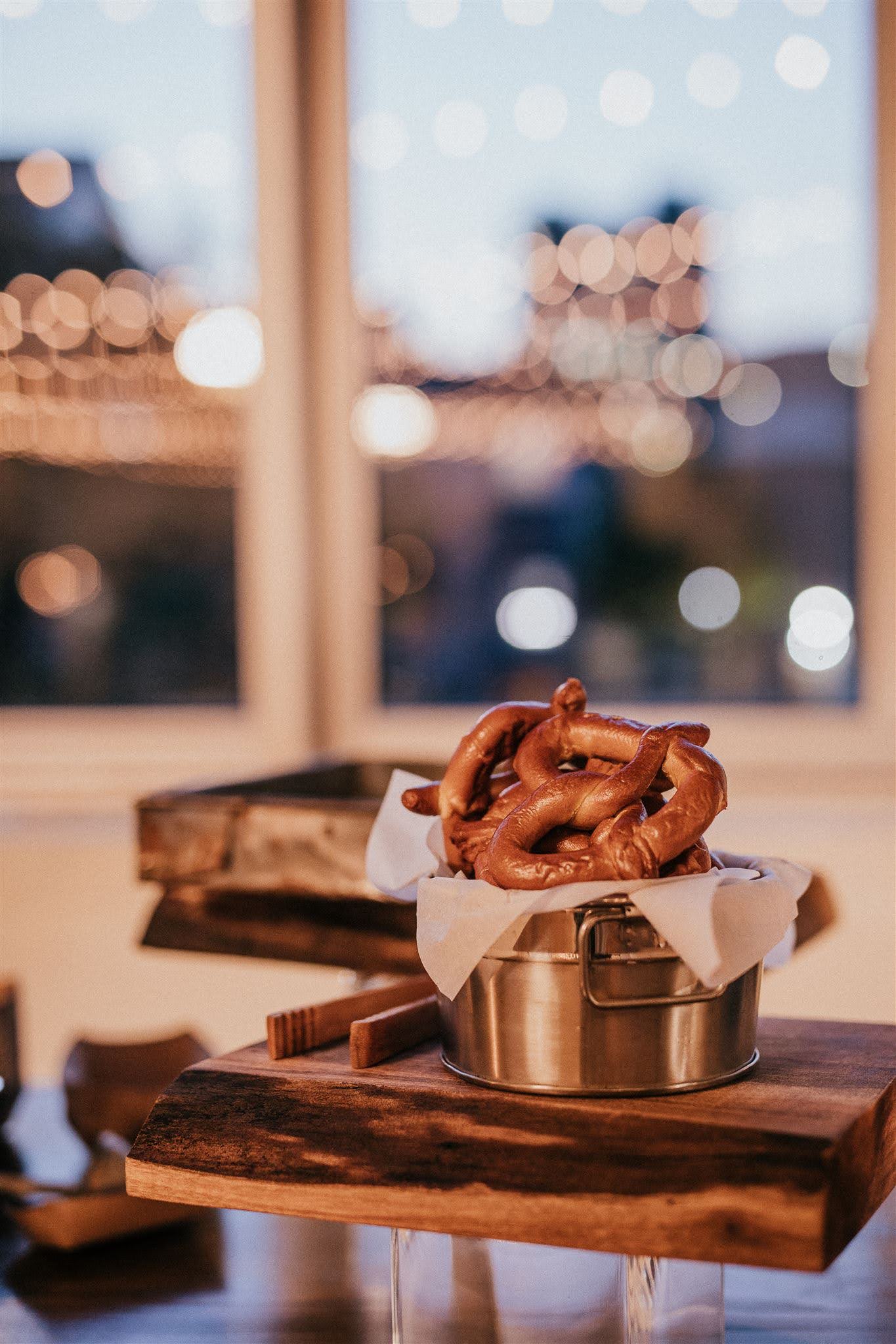 Pretzel Station—Late Night Snack Soft pretzels arranged in a metal basket on a wooden platform with tongs beside them, city lights glowing softly through nearby windows in the background.