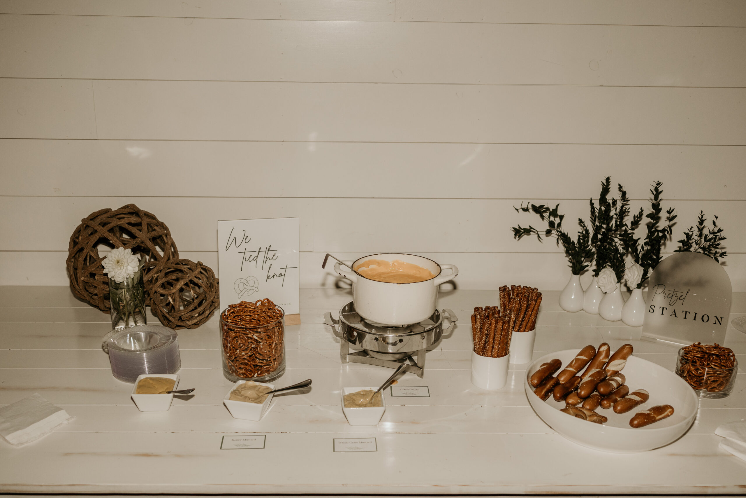 We Tied the Knot Pretzel Station— Late Night Snack The “We Tied the Knot” late-night pretzel station features soft and hard pretzel rods, warm cheese sauce, and a trio of mustards, complemented by rustic table decor including wooden balls and greenery in white vases.