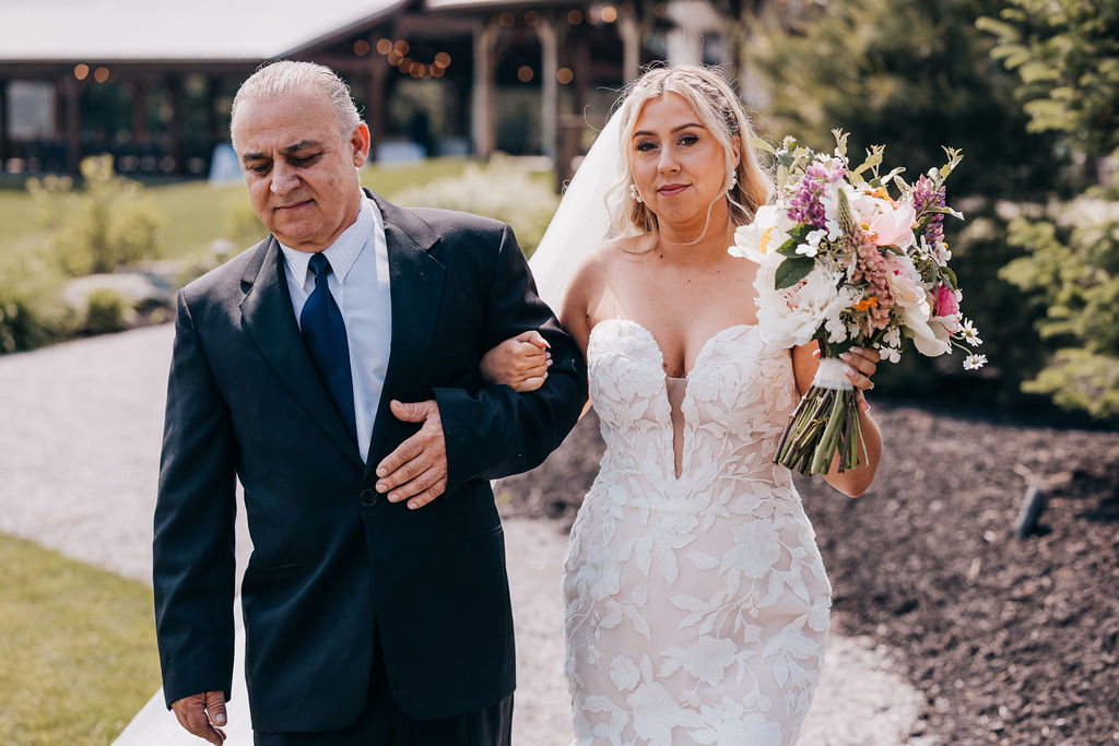 Bride aisle walk Bride in floral lace gown holding a colorful bouquet, walking down the aisle arm-in-arm with her father.