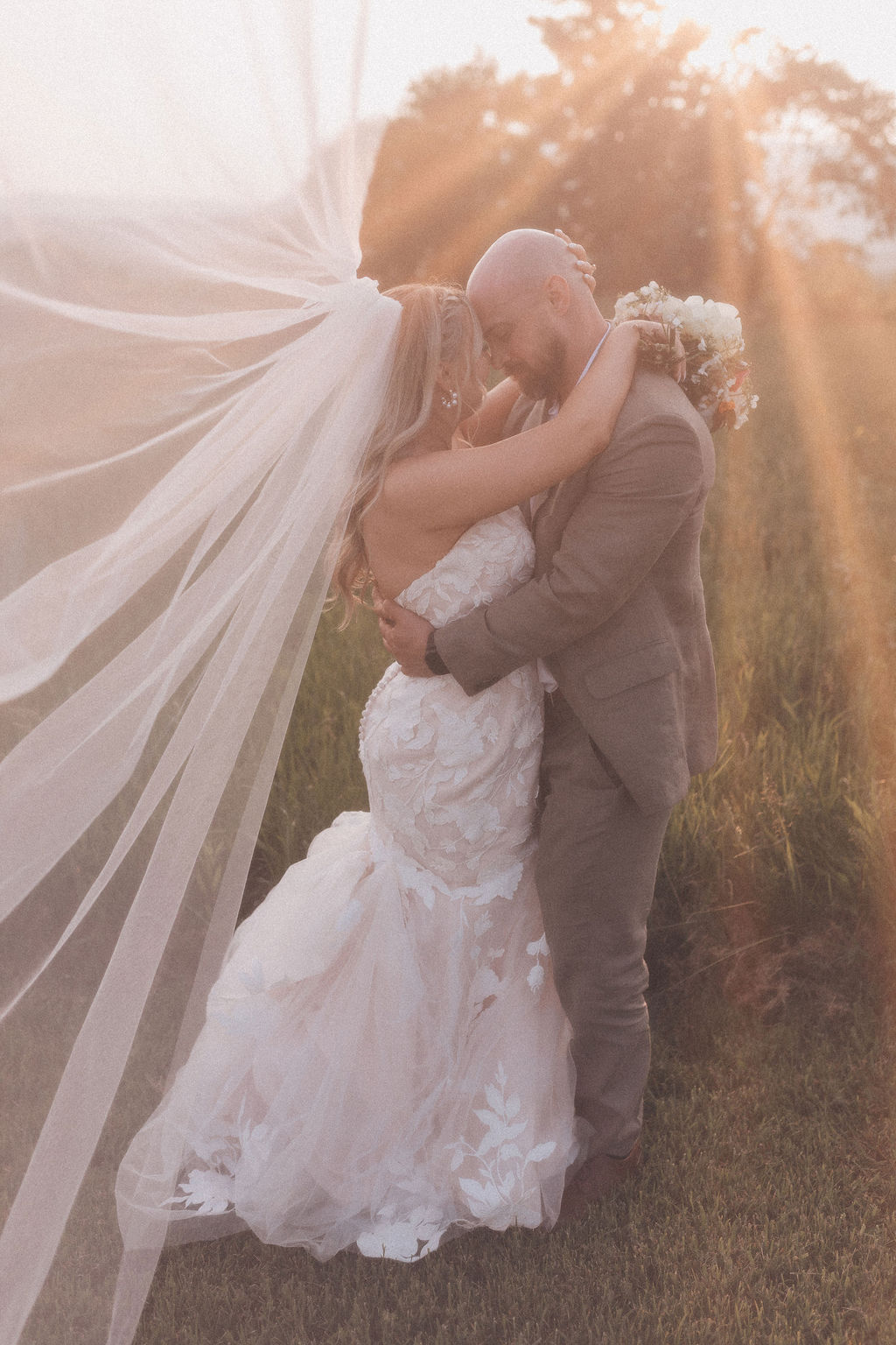 Golden Hour Wedding Embrace Bride and groom embrace in a golden field as sunlight streams through the bride’s flowing veil, creating a dreamy, romantic glow.