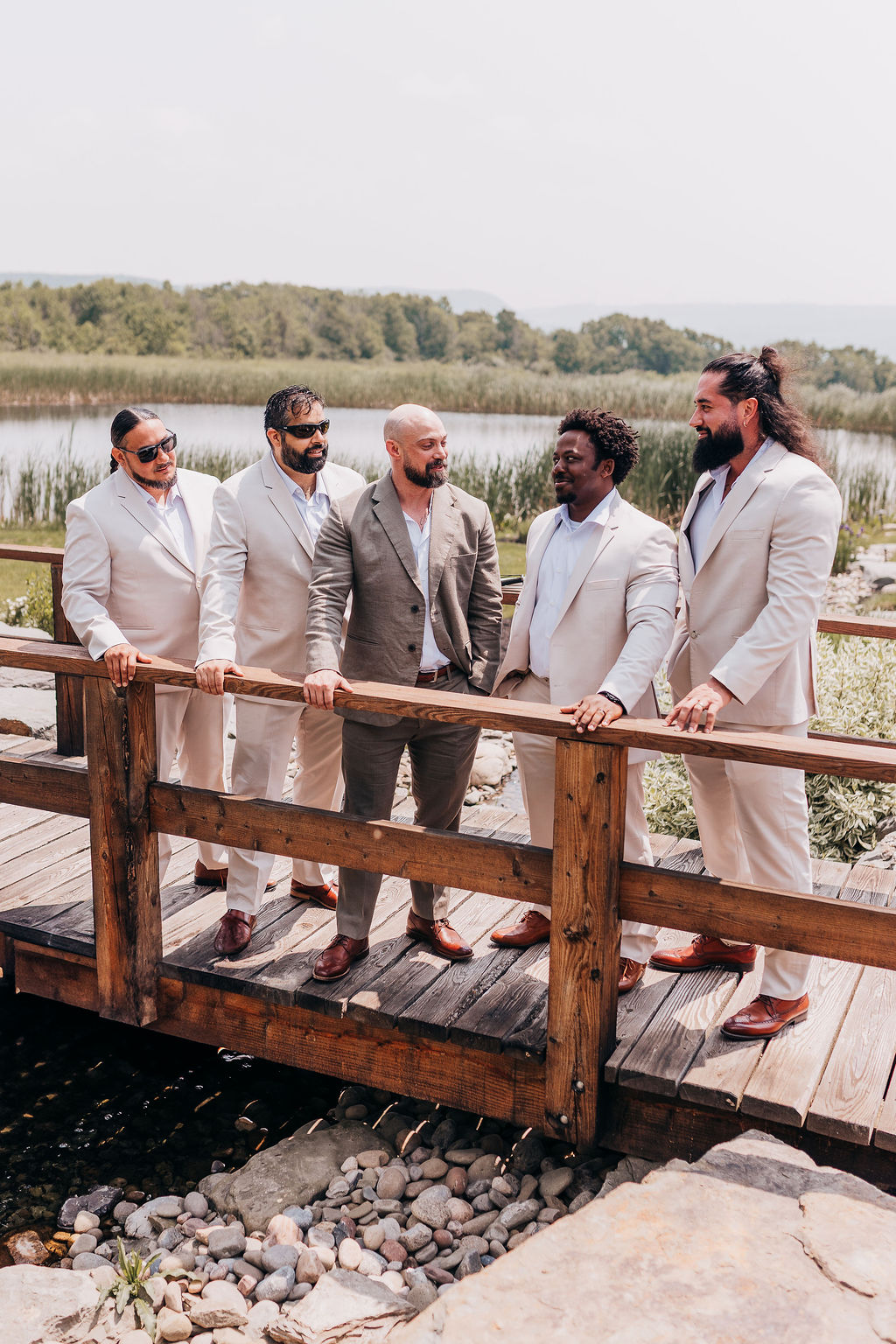 Groomsmen on the Bridge The groom and his groomsmen stand on a rustic wooden bridge over a creek, dressed in tan suits and laughing together before the ceremony.