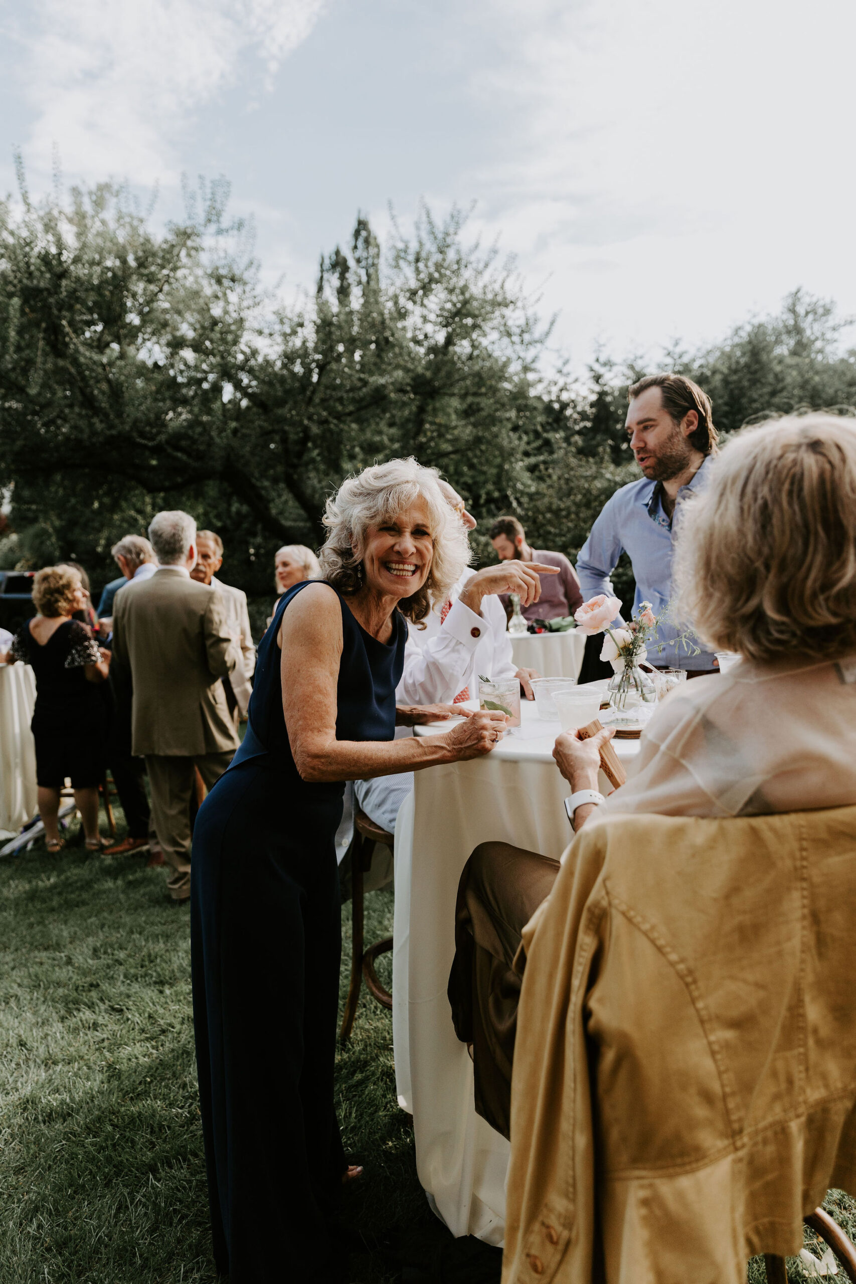 Joyful Guest at Outdoor Cocktail Party A smiling older woman in a navy jumpsuit chats with other guests around a cocktail table at an outdoor garden party.