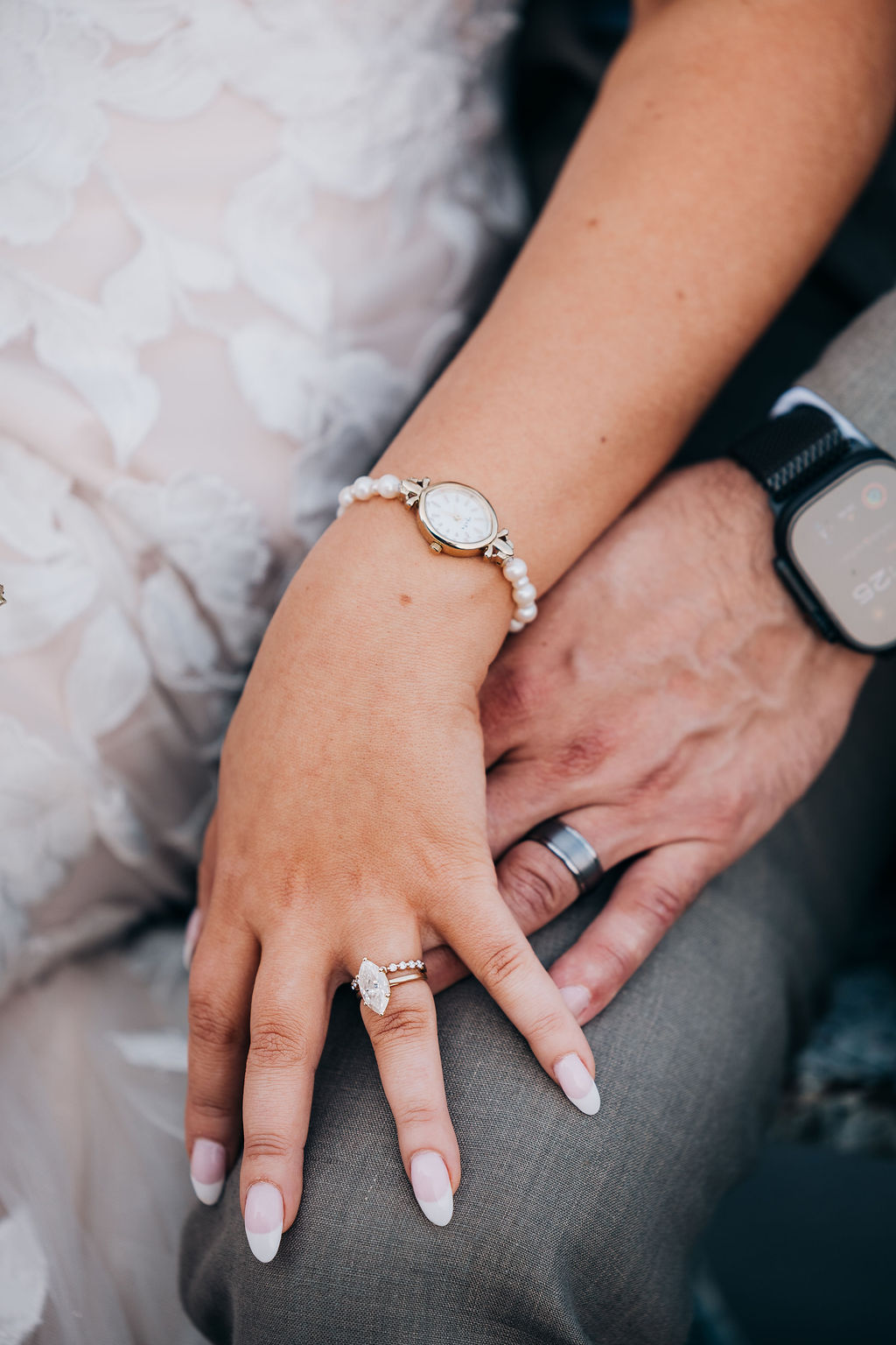 Hands with Rings The couple’s hands resting together, showcasing the bride’s diamond engagement ring, wedding band, and pearl bracelet alongside the groom’s sleek band and watch.