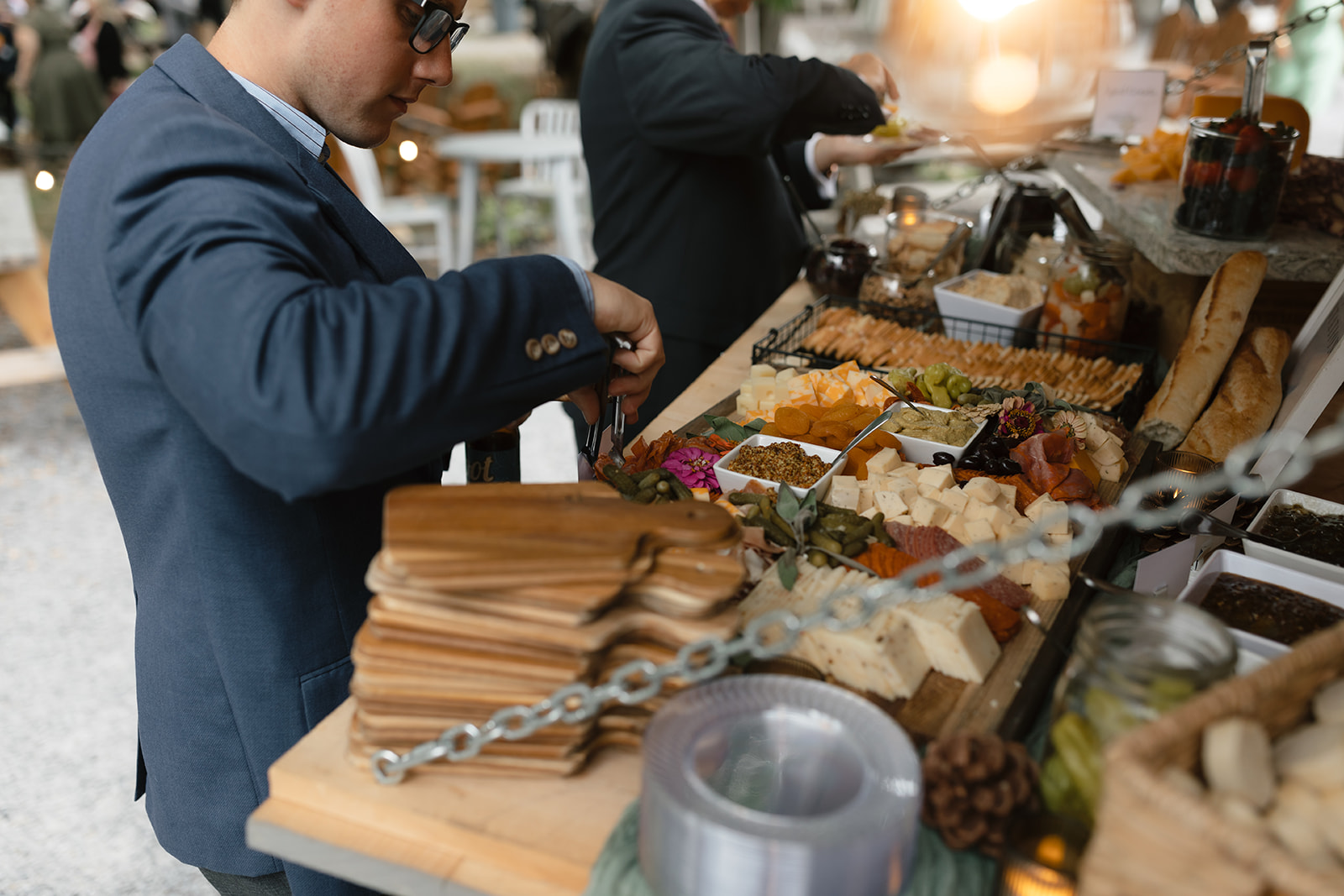 Guest Serving from Charcuterie Display A man in a suit selects from a charcuterie display with cheeses, meats, crackers, and garnishes arranged on a rustic wooden table.