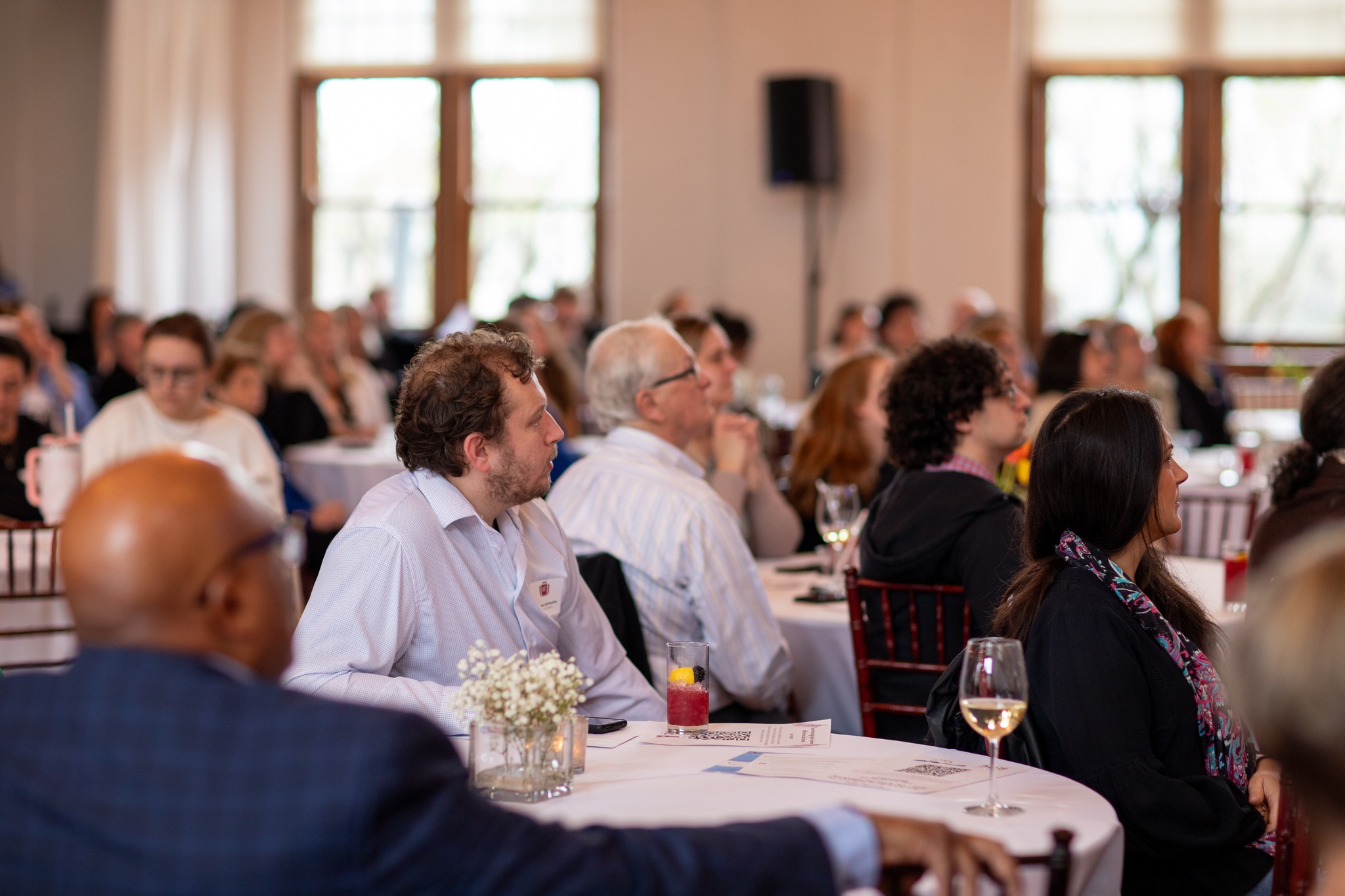 VIC – Corporate Meeting Attendees seated at round tables during a corporate meeting, attentively listening to a presentation in a bright room with large windows. Glasses of wine and floral centerpieces are on the tables.