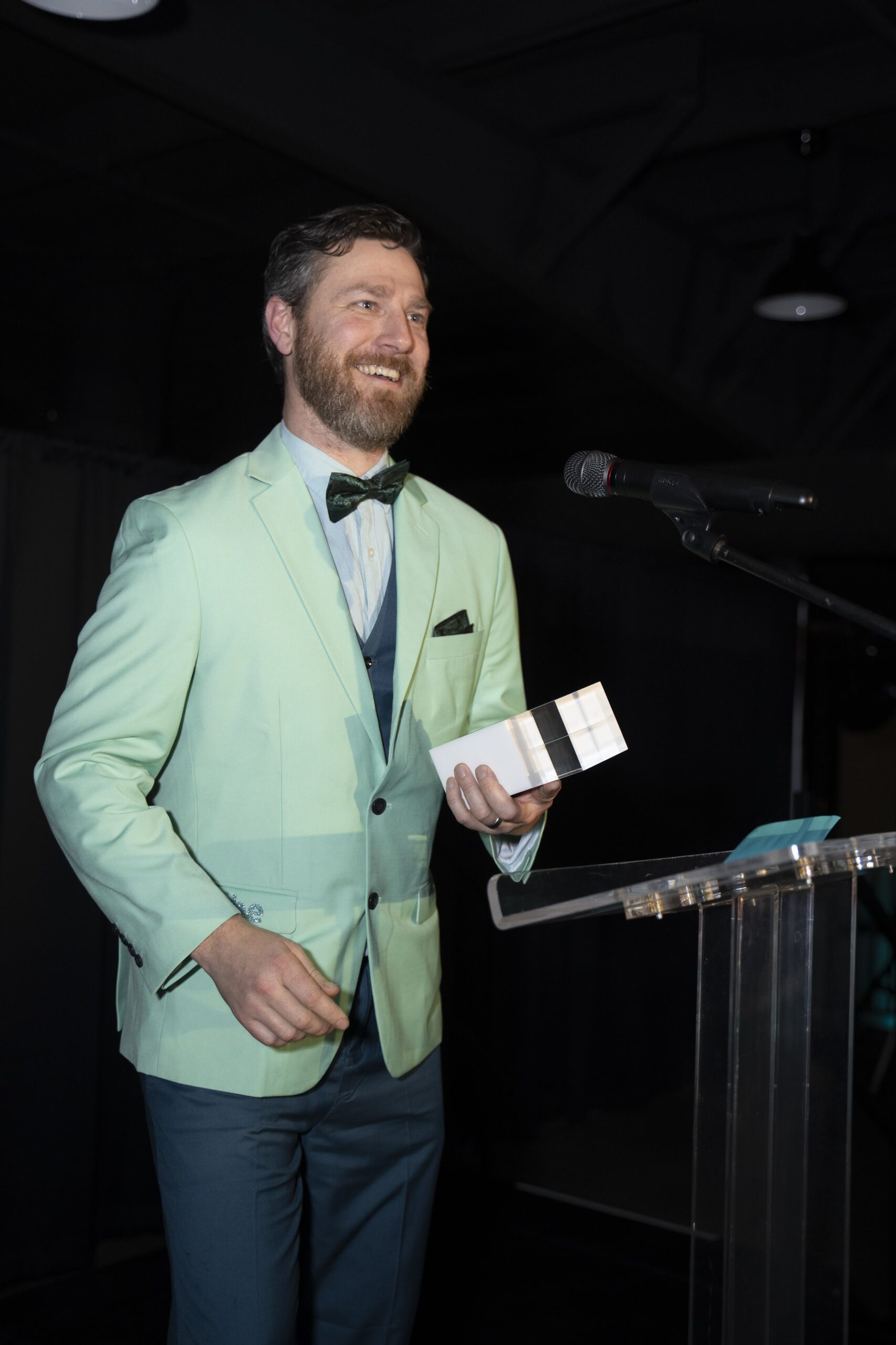 Award Acceptance at 2023 MIBE Awards Ceremony Man in a mint green suit jacket smiles while holding an award on stage at the 2023 MIBE Awards ceremony, standing behind a podium with a microphone.