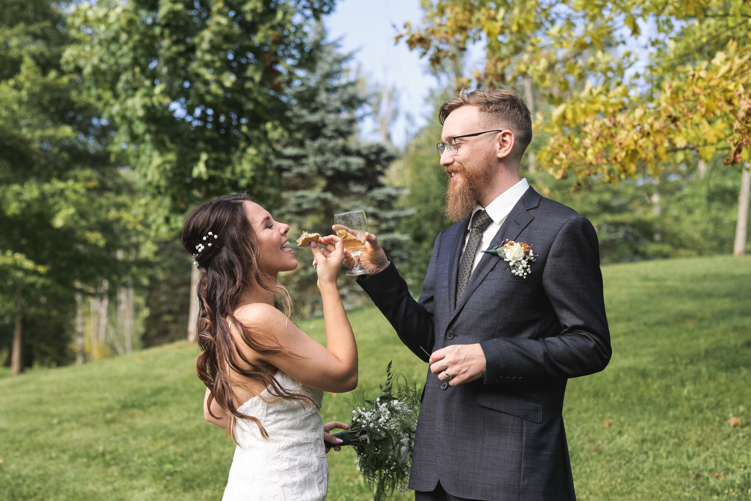 Bride and Groom Sharing a Toast Outdoors on Wedding Day A bride in a strapless lace gown and a groom in a dark suit share a playful toast and bite to eat during their outdoor wedding celebration, surrounded by lush greenery.