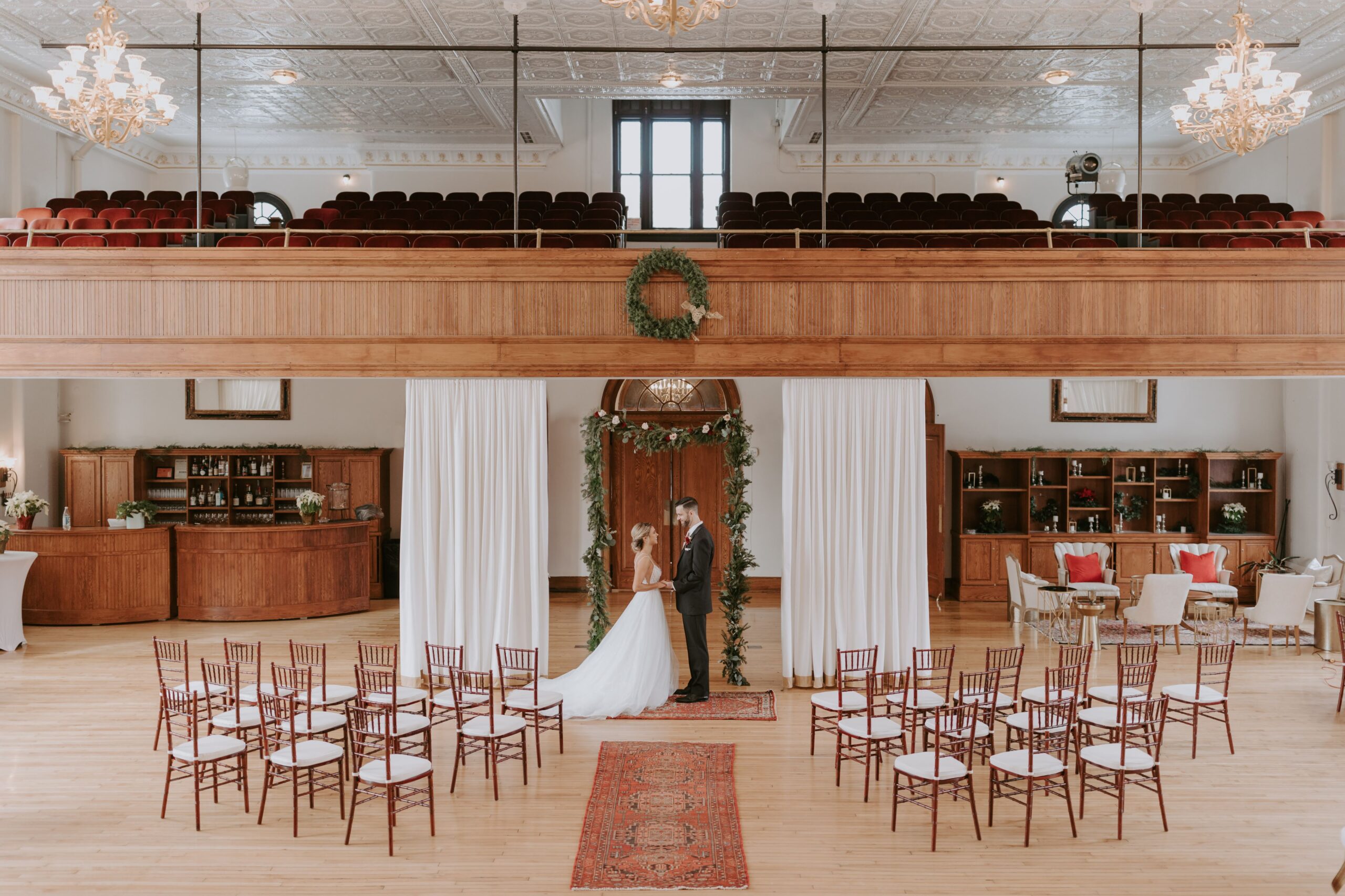 Elegant Indoor Wedding Ceremony at The Historic German House | Romantic Winter-Inspired Setting Bride and groom stand beneath a greenery-draped arch inside The Historic German House, surrounded by soft white drapery, vintage wood accents, and elegant red seating for a timeless indoor wedding ceremony.