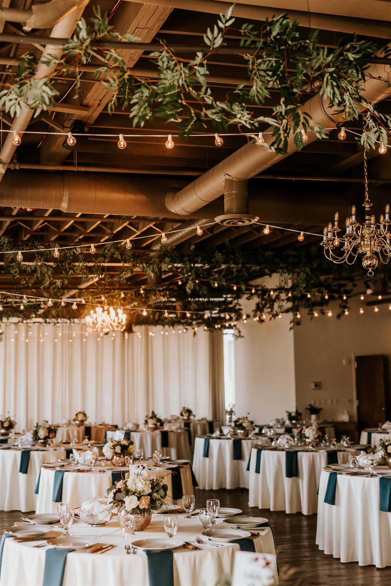 Elegant Wedding Reception Setup at Arbor Loft with Greenery and Chandeliers Arbor Loft decorated with round tables dressed in ivory linens, navy napkins, floral centerpieces, and overhead greenery with chandeliers and string lights.