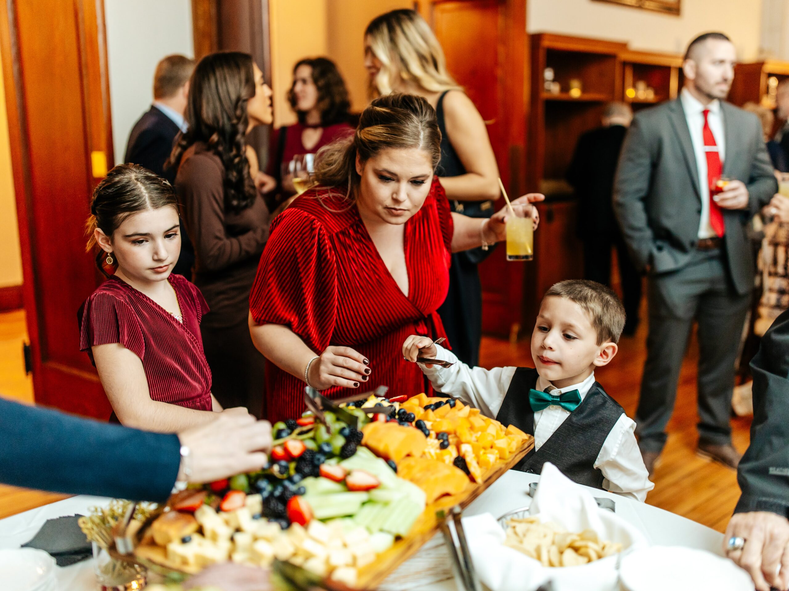 Children and adults serving themselves fruit and cheese from a grazing table during a wedding cocktail hour.