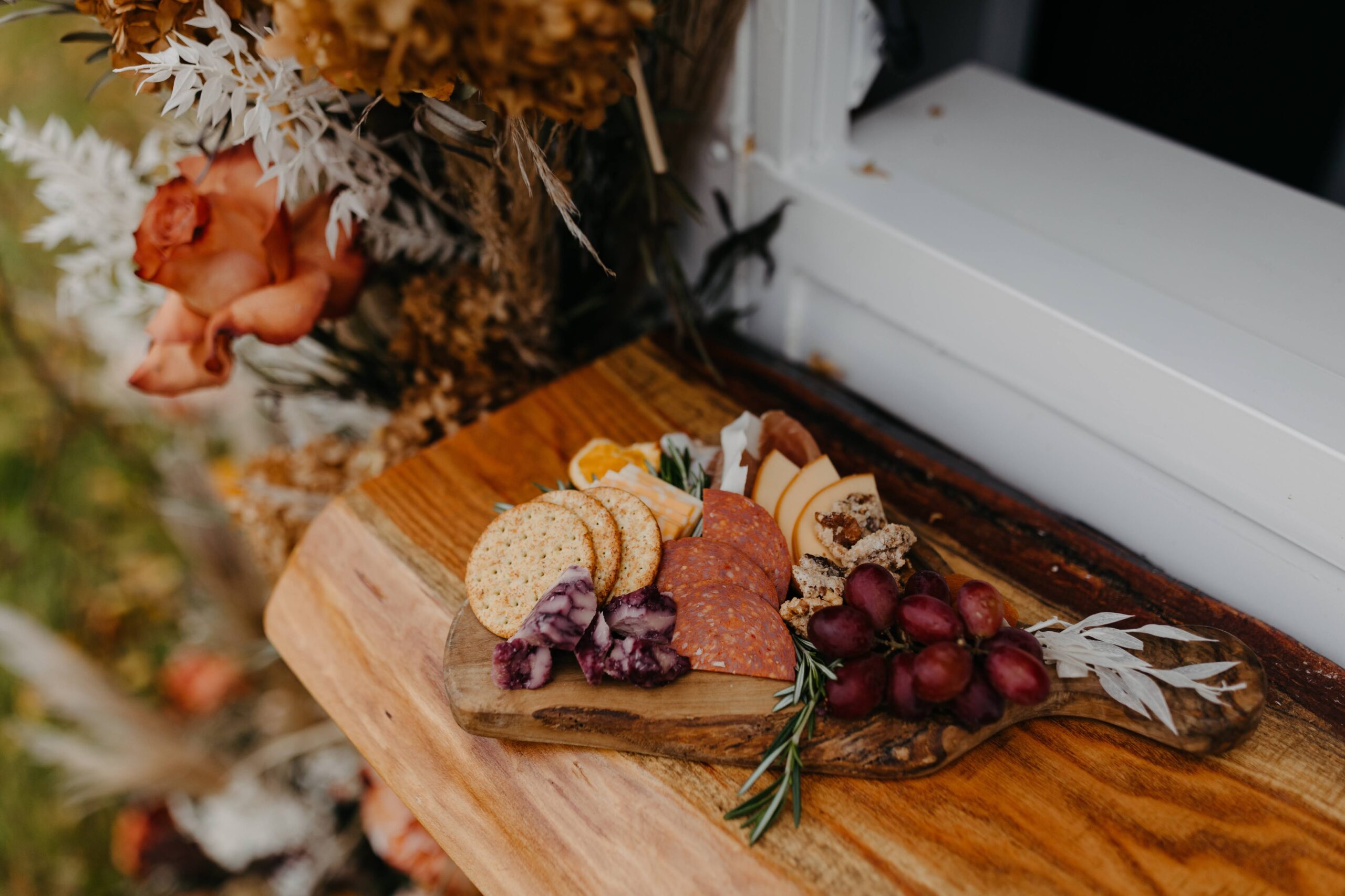 Small rustic wooden board with crackers, sliced meats, cheese, grapes, and garnishes styled with floral accents.