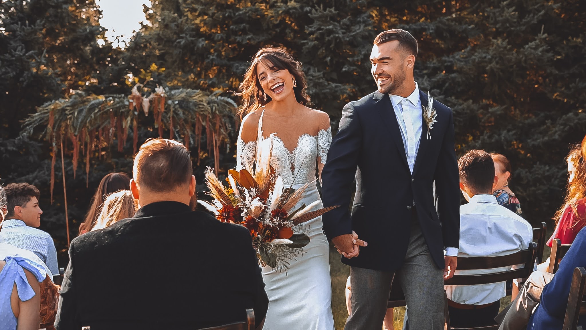 Bride and groom walking down the aisle hand-in-hand, smiling joyfully after their ceremony, with the bride holding a boho-inspired bouquet of pampas grass and warm-toned florals.