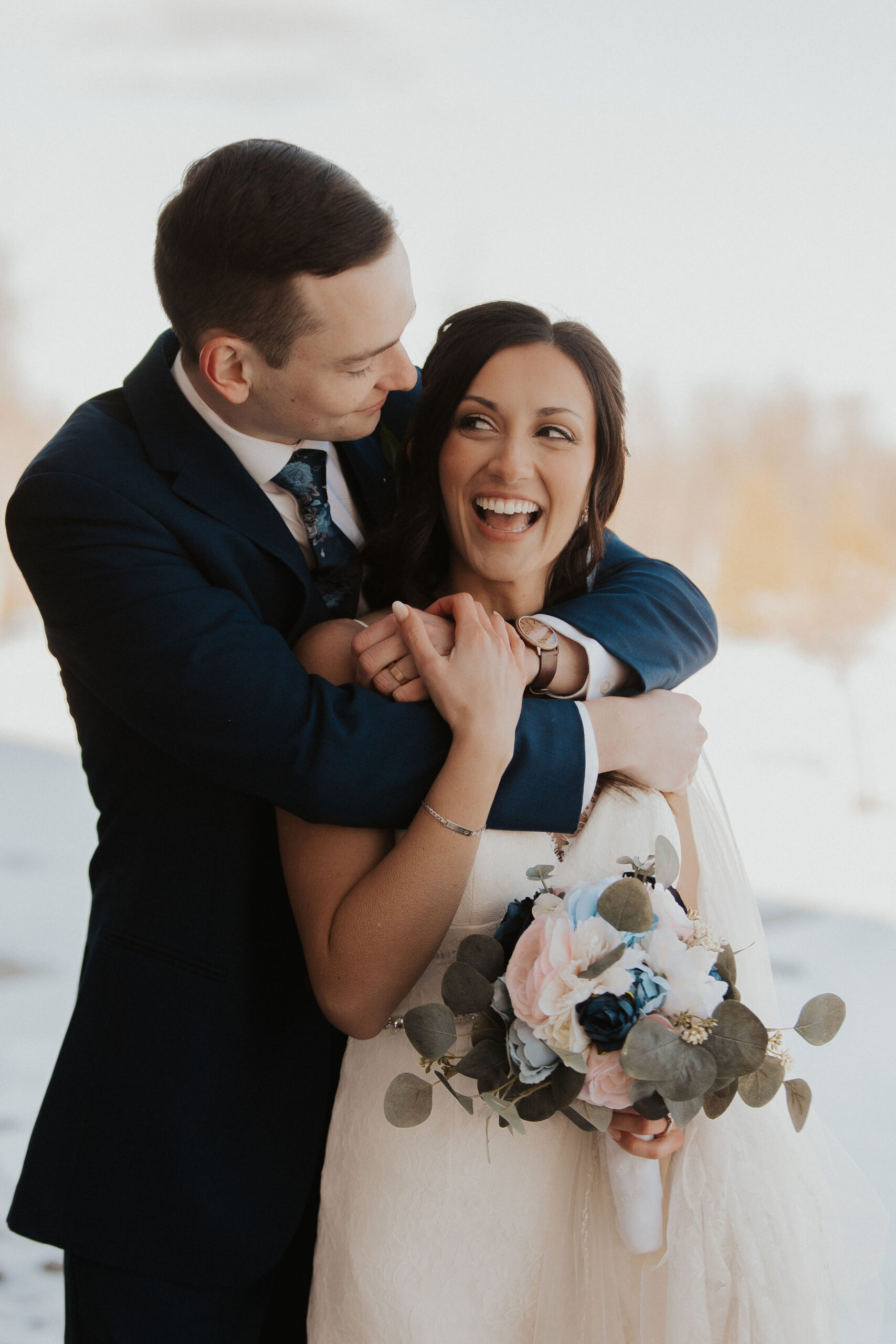 Joyful Winter Wedding | Romantic Bride and Groom Portrait Groom embraces his smiling bride during a cozy winter wedding. The bride holds a pastel bouquet with blue and blush flowers while laughing warmly, surrounded by soft snow and golden light.