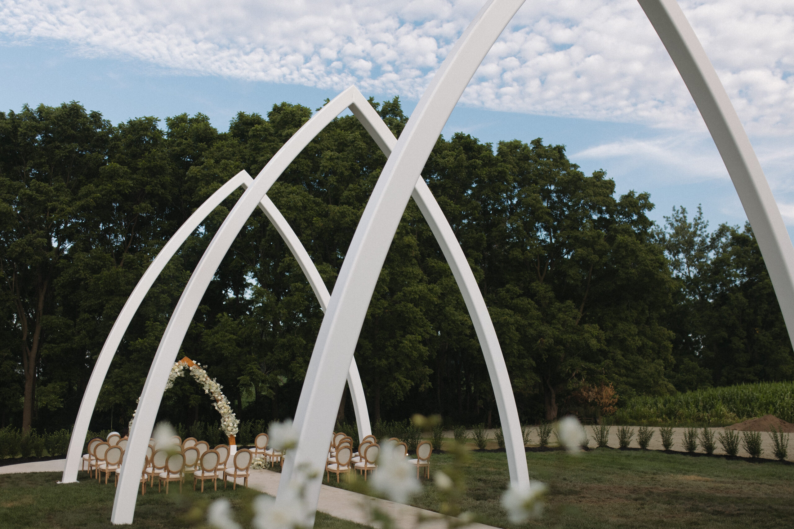 Modern Outdoor Wedding Under The Grand Arches | The Wells Estate Close-up angle of The Wells Estate’s white cathedral-style arches, set for a summer wedding ceremony with floral décor and round-back chairs against a backdrop of trees and a soft evening sky.