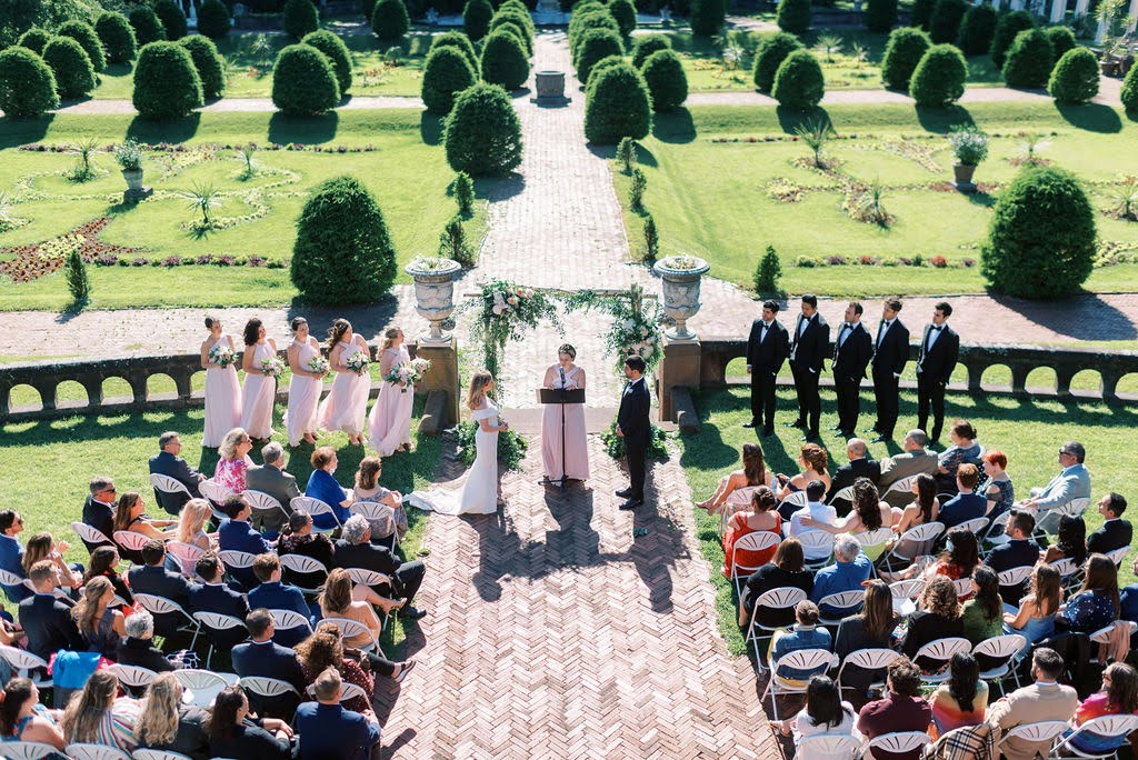 Bride and groom exchange vows under a floral arch during an outdoor wedding ceremony at Sonnenberg Gardens in Canandaigua, NY, with bridesmaids in blush dresses, groomsmen in black suits, and guests seated in a formal garden setting.