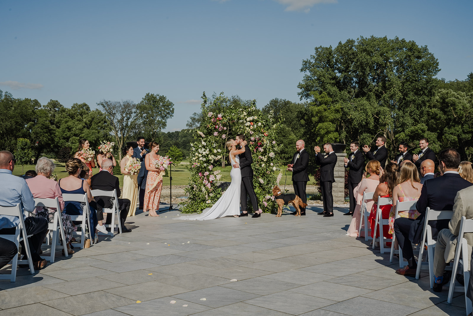 Bride and groom share their first kiss under a lush floral arch during an outdoor wedding ceremony at The Annex, surrounded by bridesmaids, groomsmen, guests, and even their dog in attendance.