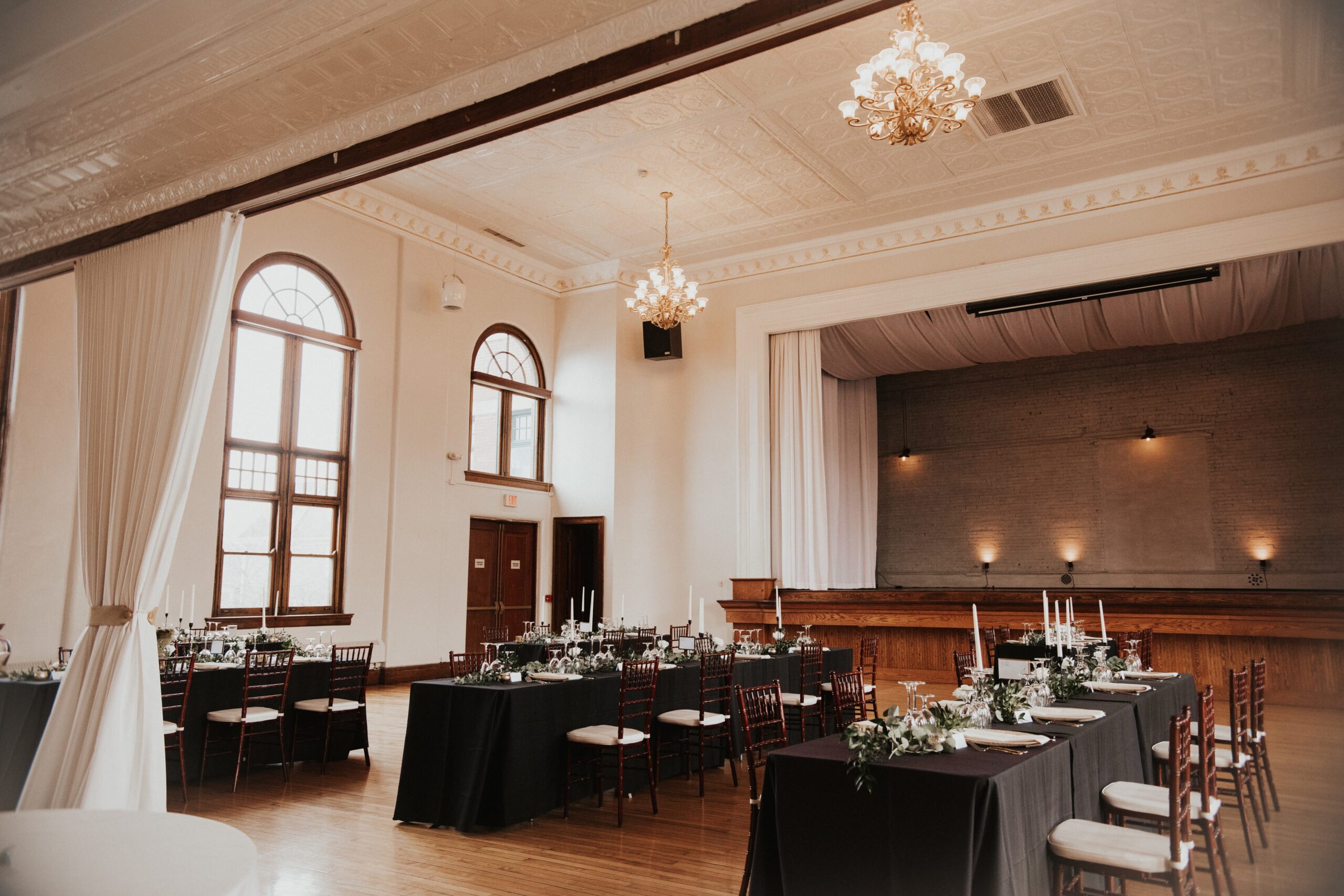 Romantic Ballroom Setup with Tall Floral Centerpieces | The Historic German House Wide view of The Historic German House ballroom set for an elegant wedding reception, featuring black table linens, greenery, and natural light through arched windows.