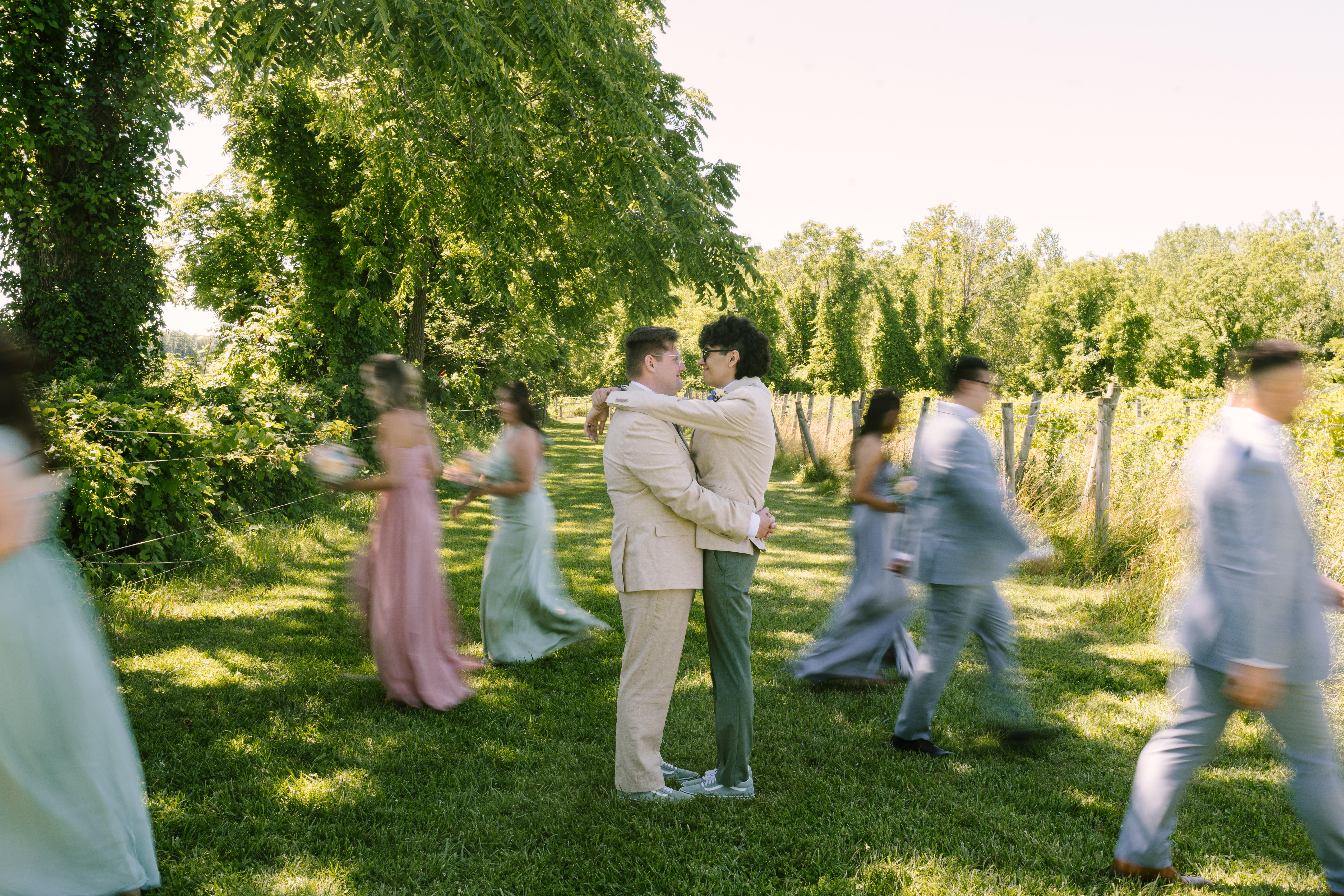 Newlyweds embrace in a vineyard path surrounded by their wedding party moving around them, blending motion and stillness in a dreamy outdoor portrait at Deer Run Winery.