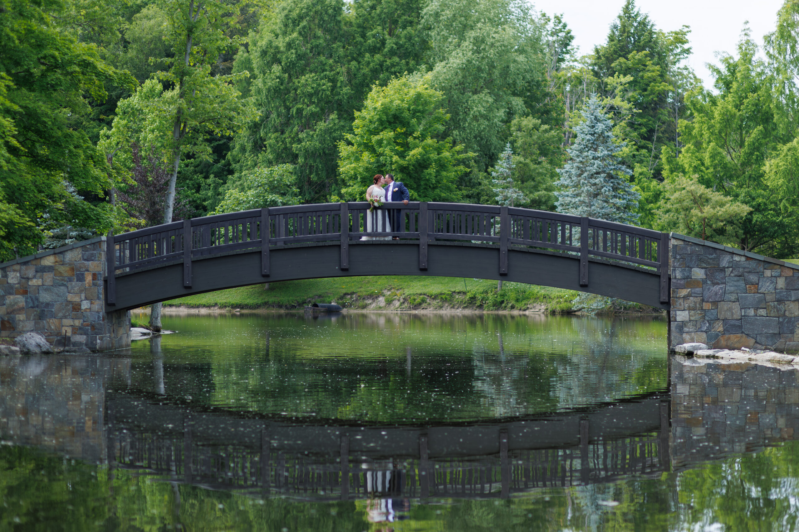 Bride and groom sharing a kiss on the stone and wood bridge over the pond at The Lodge at Shadow Hill, surrounded by lush greenery and reflections in the water.