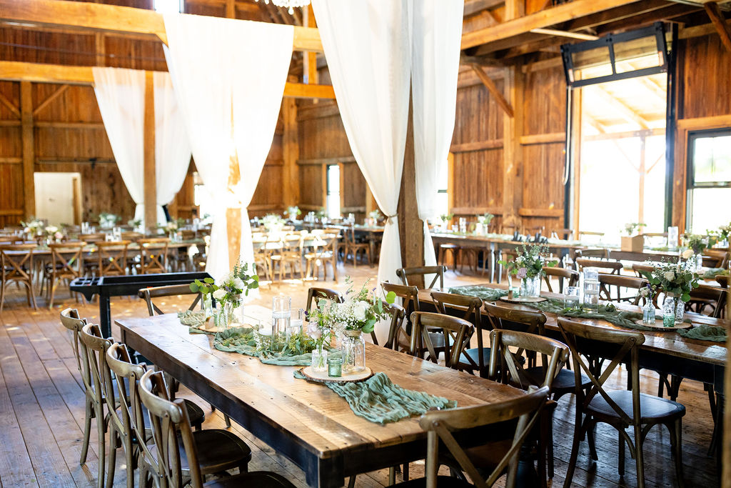 Rustic Barn Wedding Reception at Graceful Gatherings | Elegant Farmhouse Decor Long wooden farmhouse tables with greenery runners, simple floral centerpieces, and white draped fabric inside a rustic barn wedding venue at Graceful Gatherings in New York.