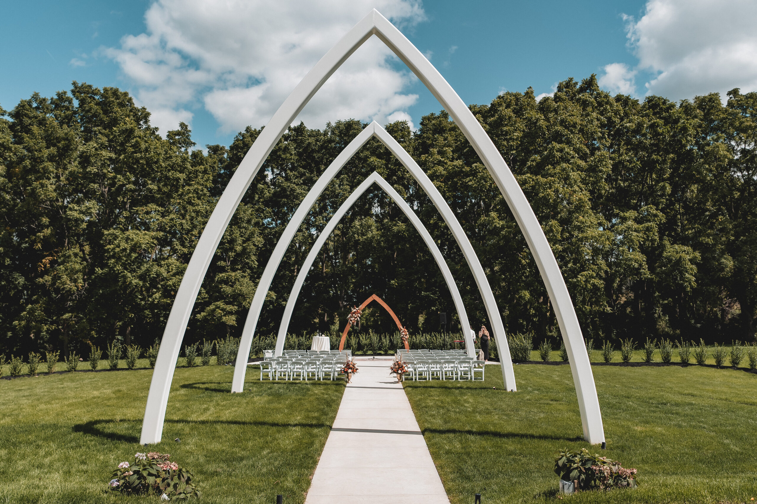 Wedding Ceremony | The Wells Estate Outdoor Arches Wide view of The Wells Estate’s Grand Arches, featuring elegant white gothic arches framing a romantic outdoor wedding ceremony under a blue sky surrounded by lush greenery.