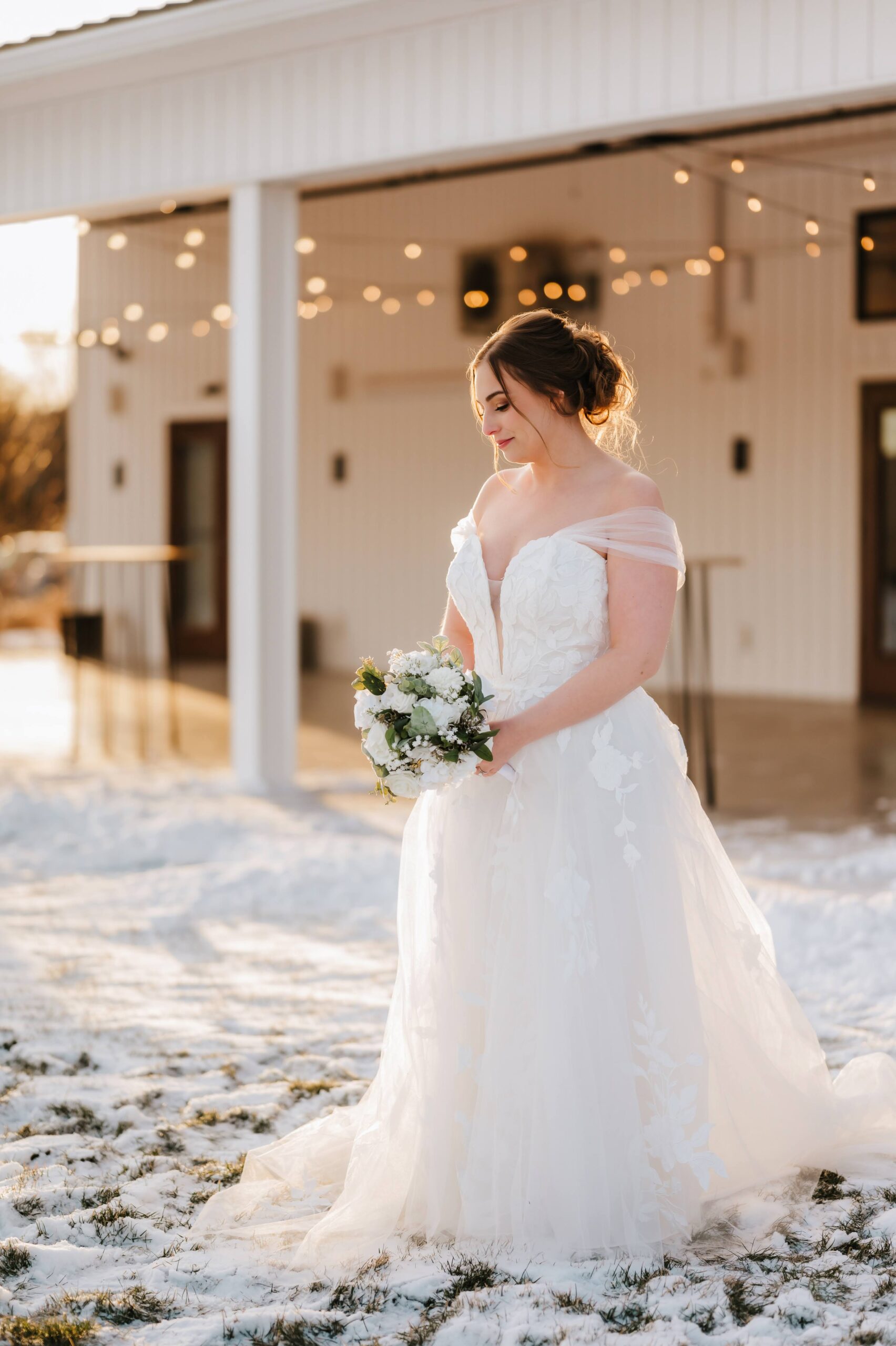 Winter Bride | Golden Hour Wedding Portrait in Snow Bride stands in soft golden hour light, wearing an off-the-shoulder lace gown and holding a white floral bouquet. Snow glistens on the ground as string lights twinkle behind her, capturing a serene winter wedding moment.