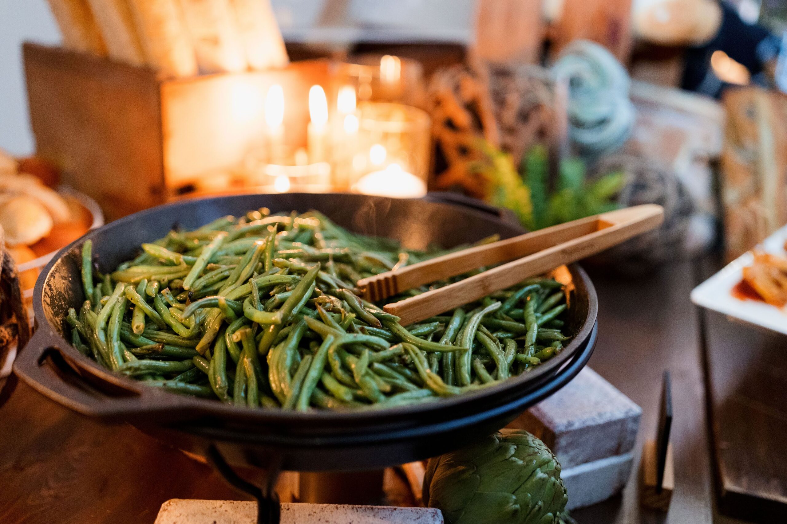 A serving dish of freshly sautéed green beans glistening with garlic butter, styled on a rustic buffet table by candlelight for an elegant catered event.