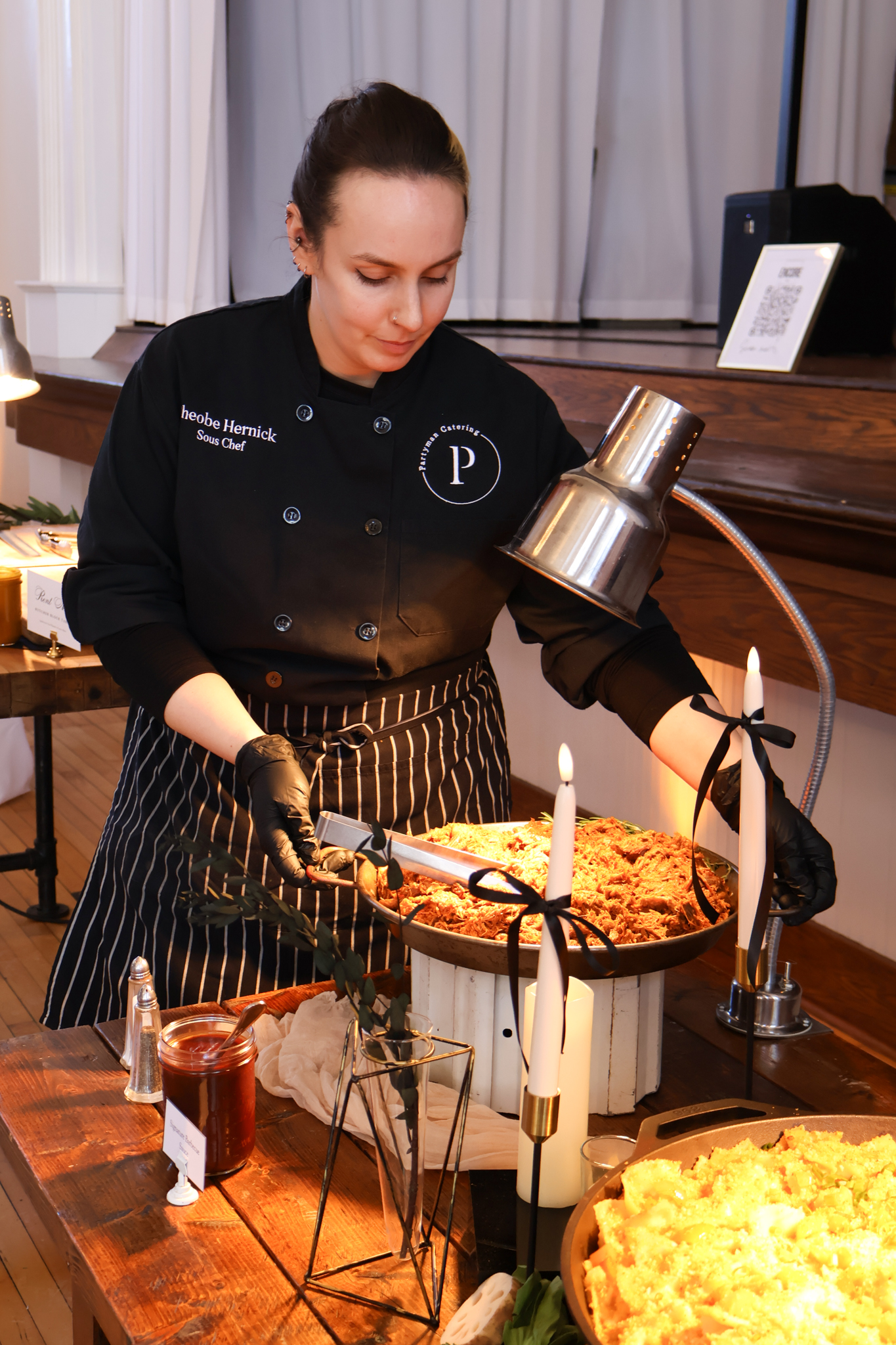 Sous Chef Phoebe Henrick of Partyman Catering serves freshly carved barbecue at a tasting event, standing behind a rustic buffet setup with glowing candles and elegant table styling.