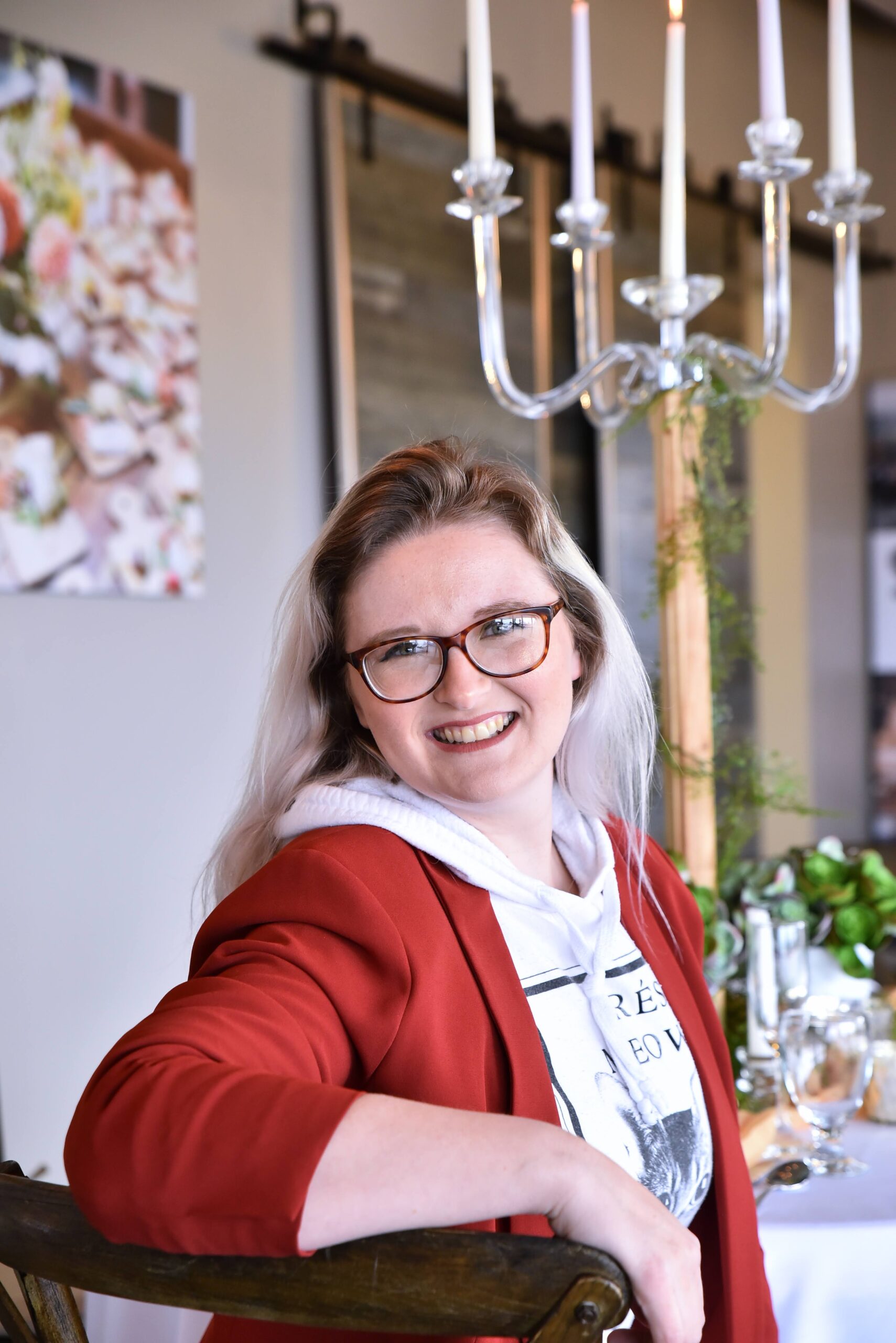 A smiling woman with glasses and a red blazer sits on a wooden chair in a beautifully set event space, with a candelabra and decorated table in the background.