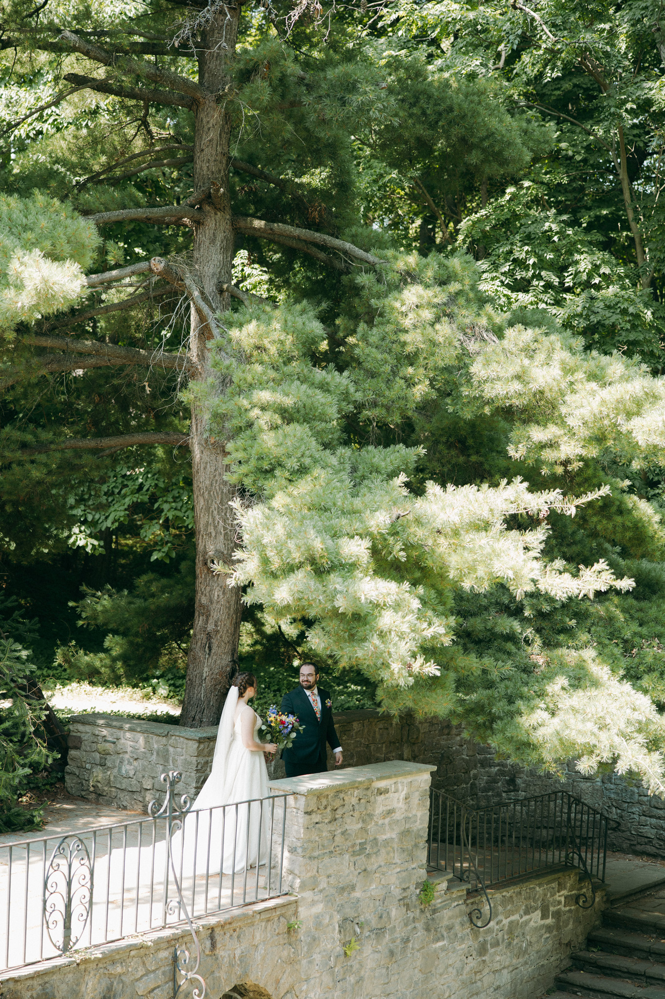 The bride and groom walk hand-in-hand along a stone garden bridge, surrounded by tall trees and soft filtered sunlight.