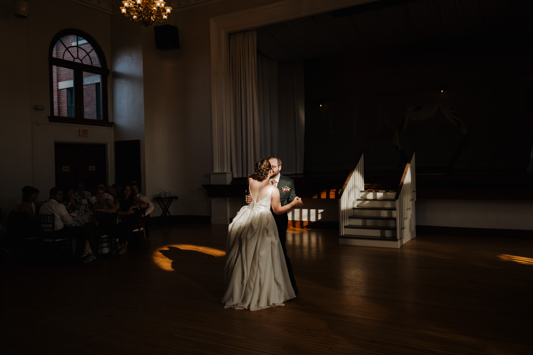 The bride and groom embrace during their first dance under dramatic lighting, with a chandelier glowing above and guests watching from the sidelines.