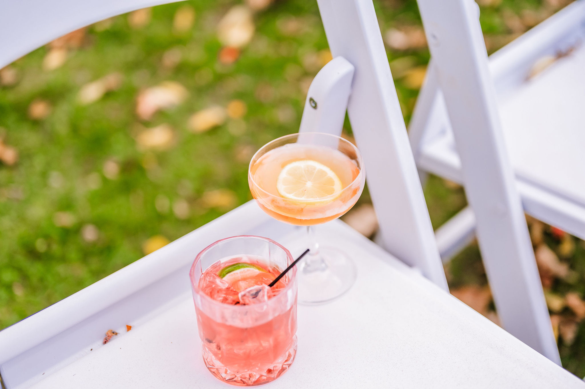 Signature Wedding Cocktails Styled on White Ceremony Chairs Pink signature cocktails in coupe and rocks glasses resting on white ceremony chairs, styled outdoors on a green lawn for a wedding celebration.