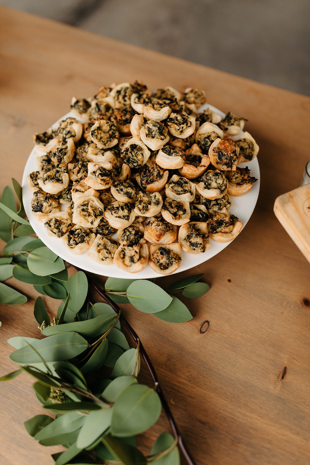 A white plate filled with golden-baked spinach puff pastry bites, displayed on a wooden table with fresh greenery, styled for an elegant event or wedding reception.