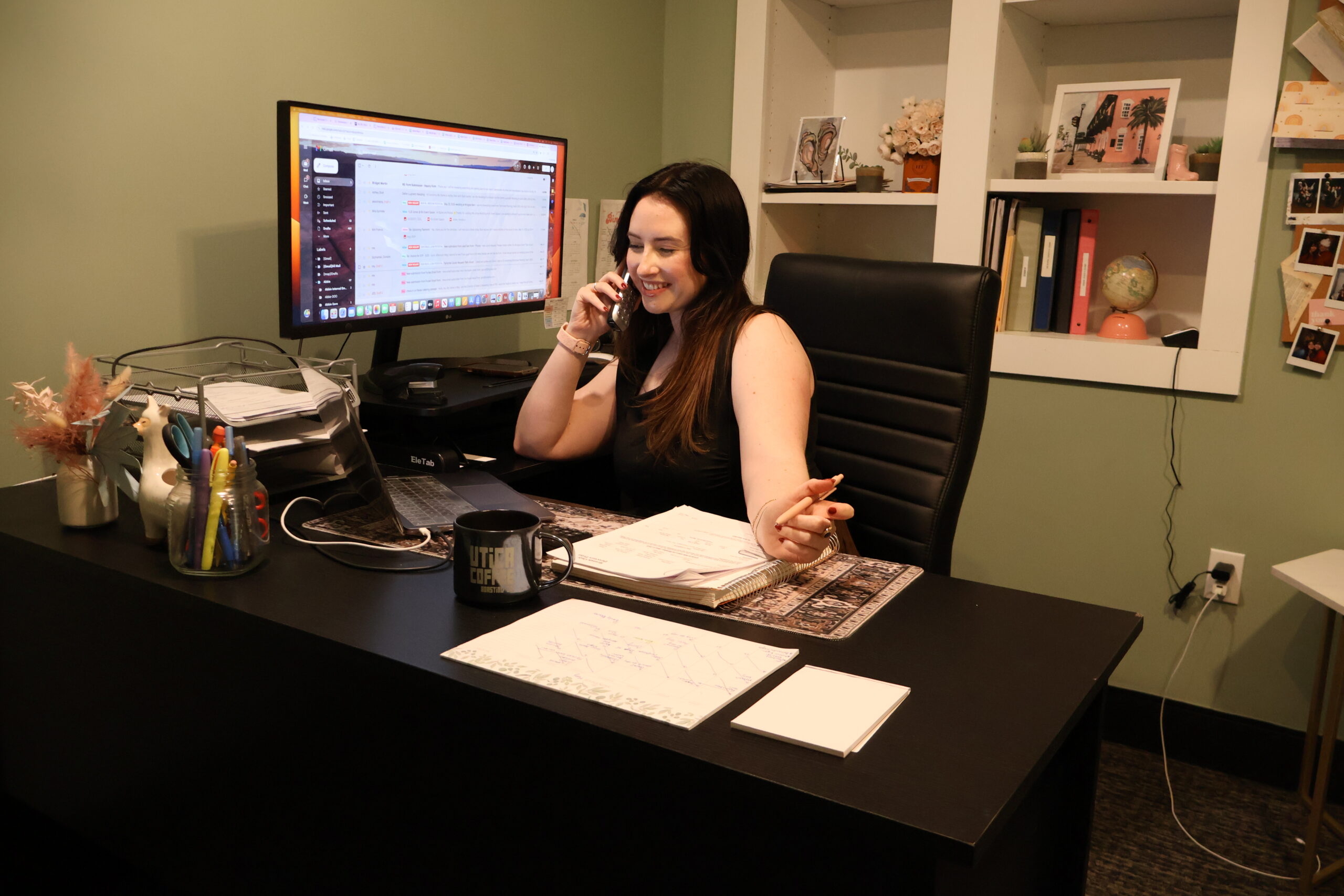 Female venue coordinator smiling while speaking on the phone at her desk, surrounded by wedding planning notes, a computer screen displaying emails, and a workspace filled with personal touches and organization tools.