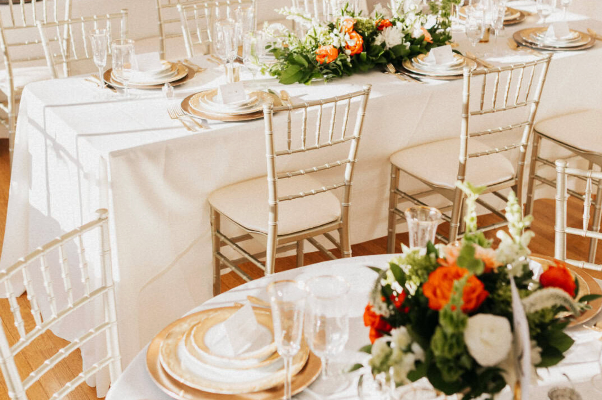 Close-up of a formal wedding table setting at Whitehall Mansion featuring gold charger plates, ivory napkins, elegant glassware, and a lush orange and white floral centerpiece.