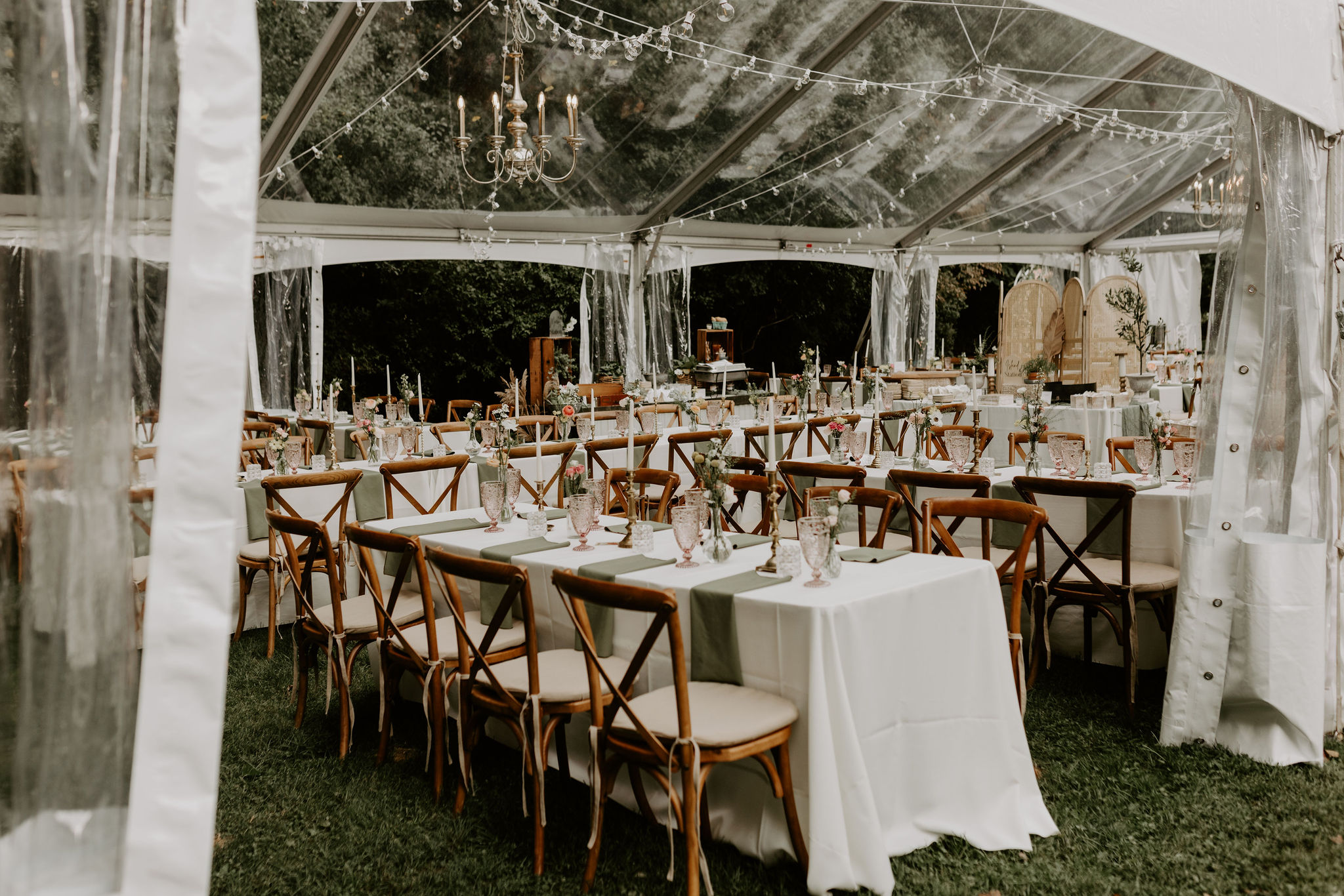 Outdoor wedding reception setup inside a clear top tent with string lights, chandelier, crossback chairs, and decorated dining tables on lawn.