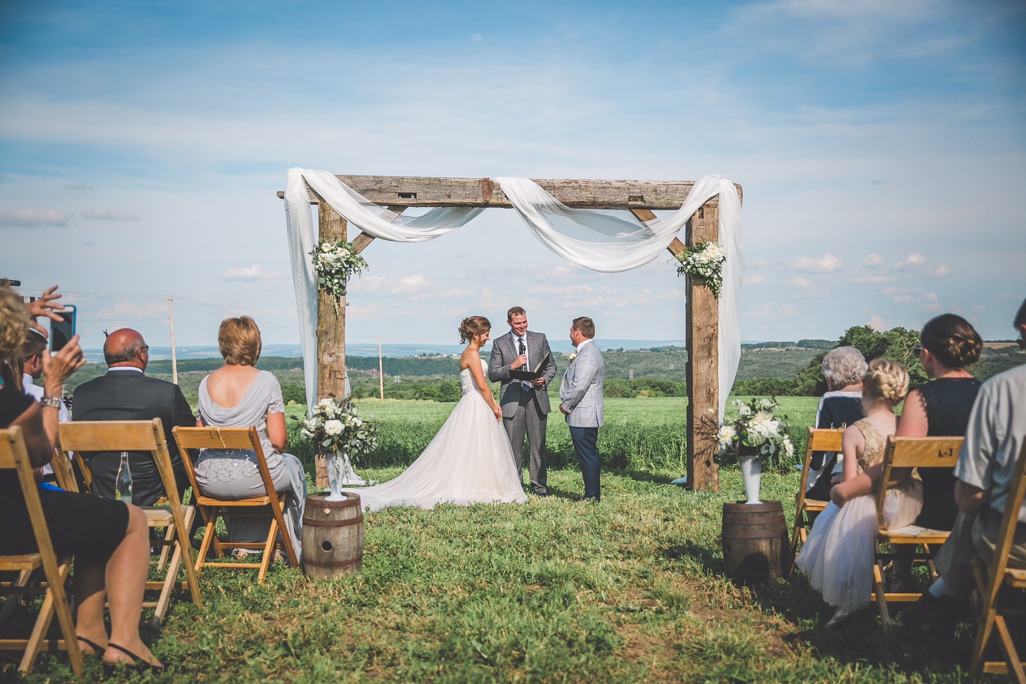 Bride and groom exchanging vows under a rustic wooden wedding arch with draped fabric during an outdoor countryside ceremony with guests seated on wooden chairs.