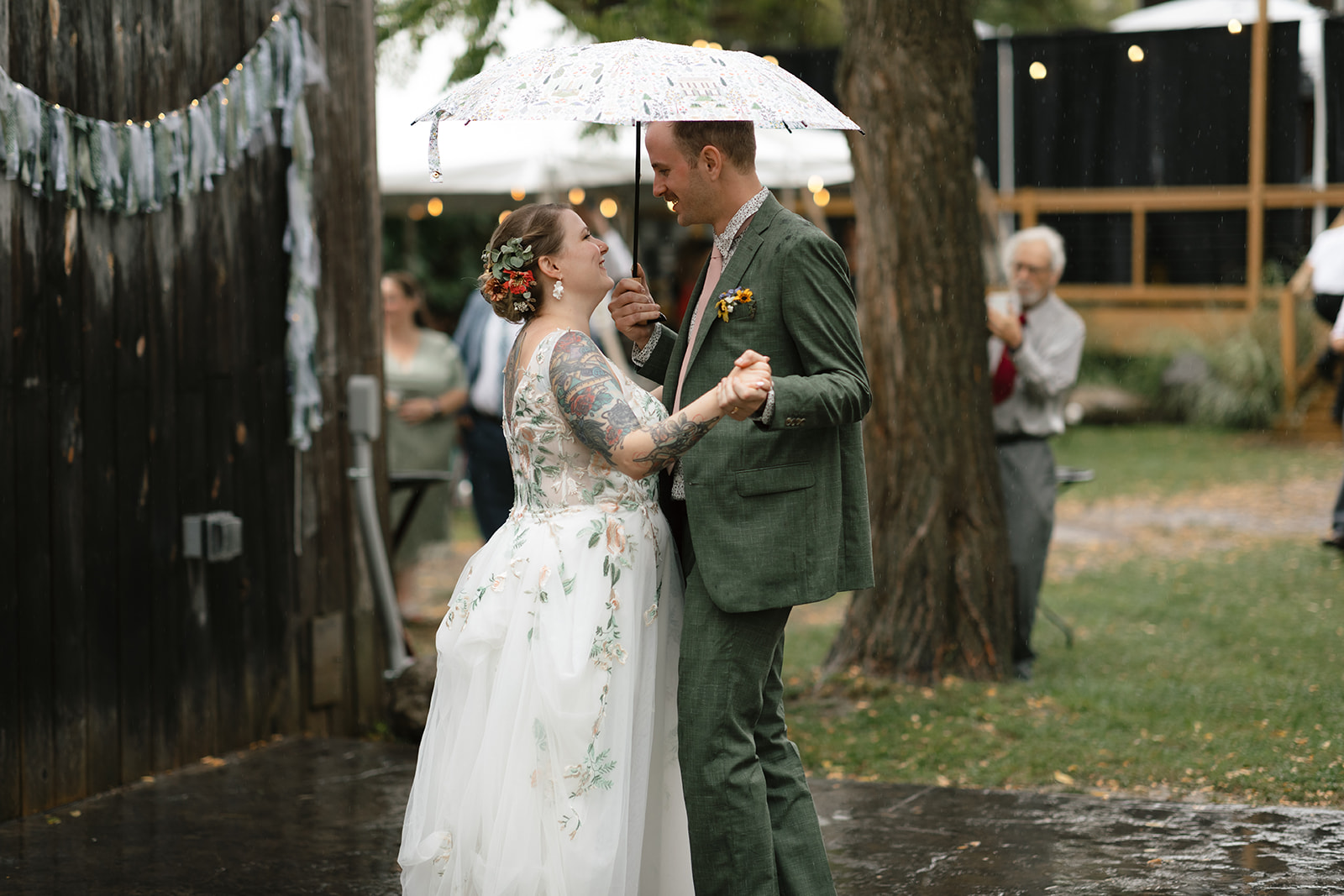 Bride and groom sharing a first dance under a patterned umbrella in the rain during an outdoor wedding reception beside a rustic barn.