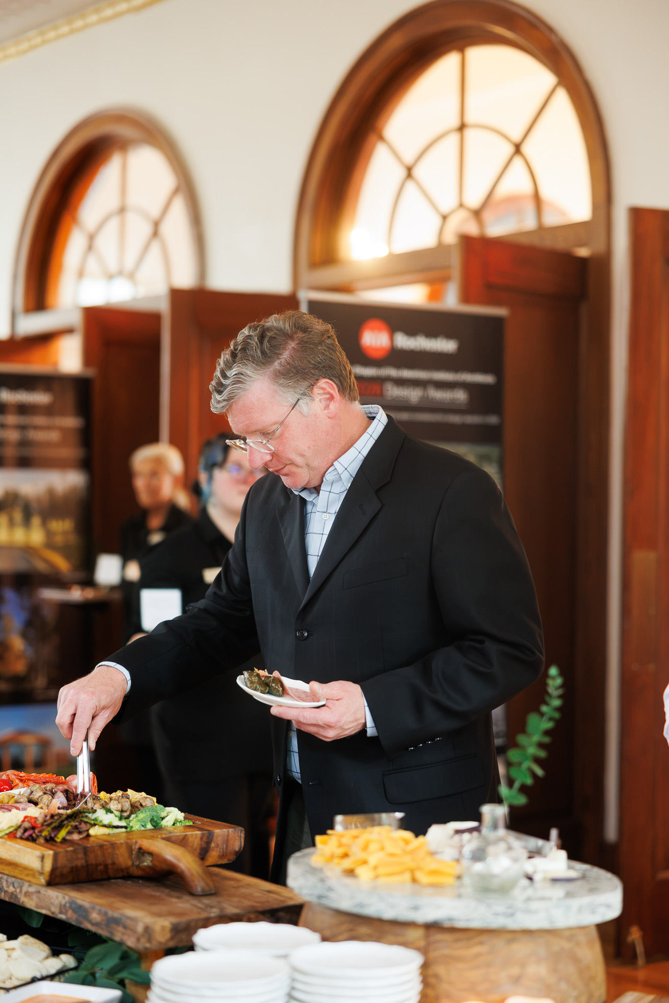 Guest Enjoying Partyman Catering Grazing Table at The Historic German House in Rochester NY Guest serving themselves from an elegant grazing table featuring artisan cheeses, charcuterie, grilled vegetables, and fresh accompaniments during a corporate event at The Historic German House in Rochester, NY.