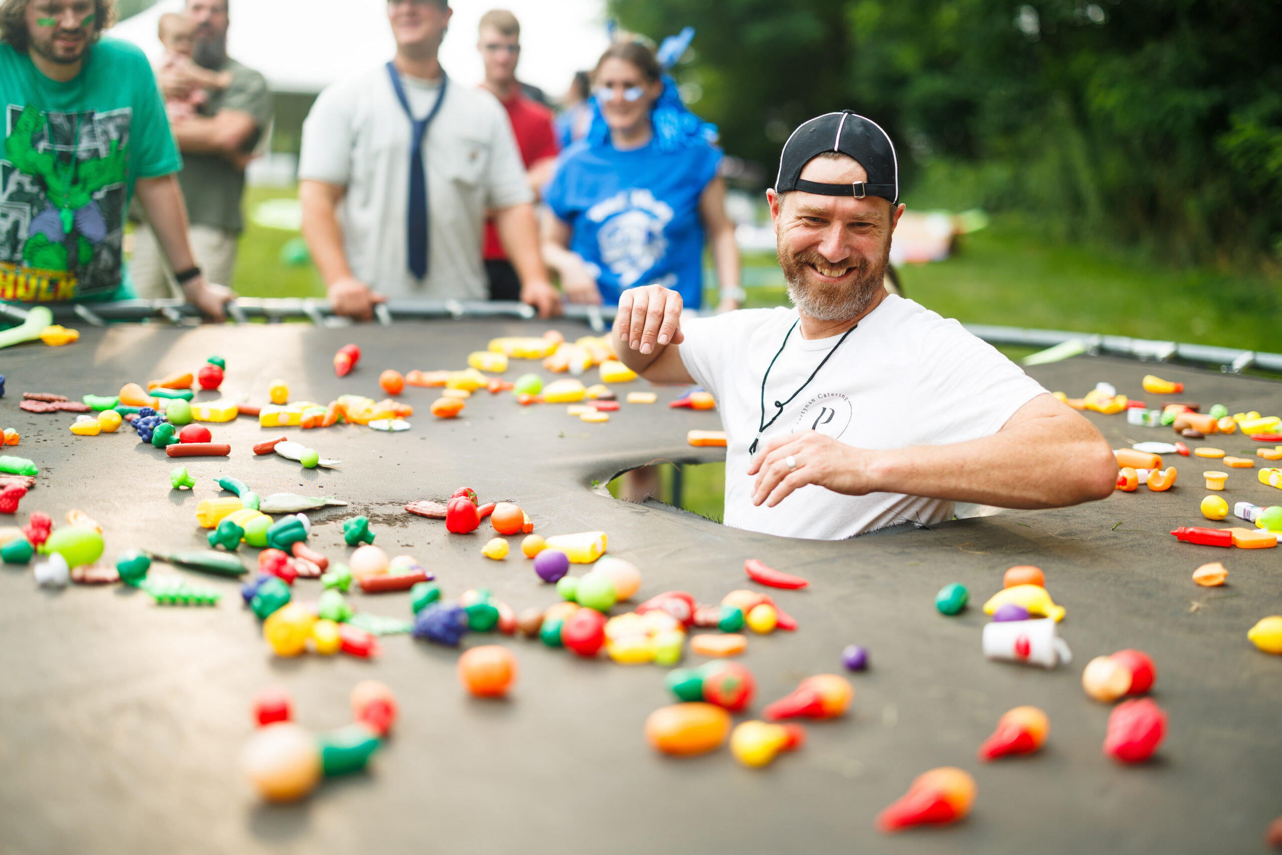 Funner Olympics Team Building Whack-A-Snacker game at Partyman Catering Partyman team member laughs while participating in a playful trampoline challenge surrounded by colorful toy food during the Funner Olympics.