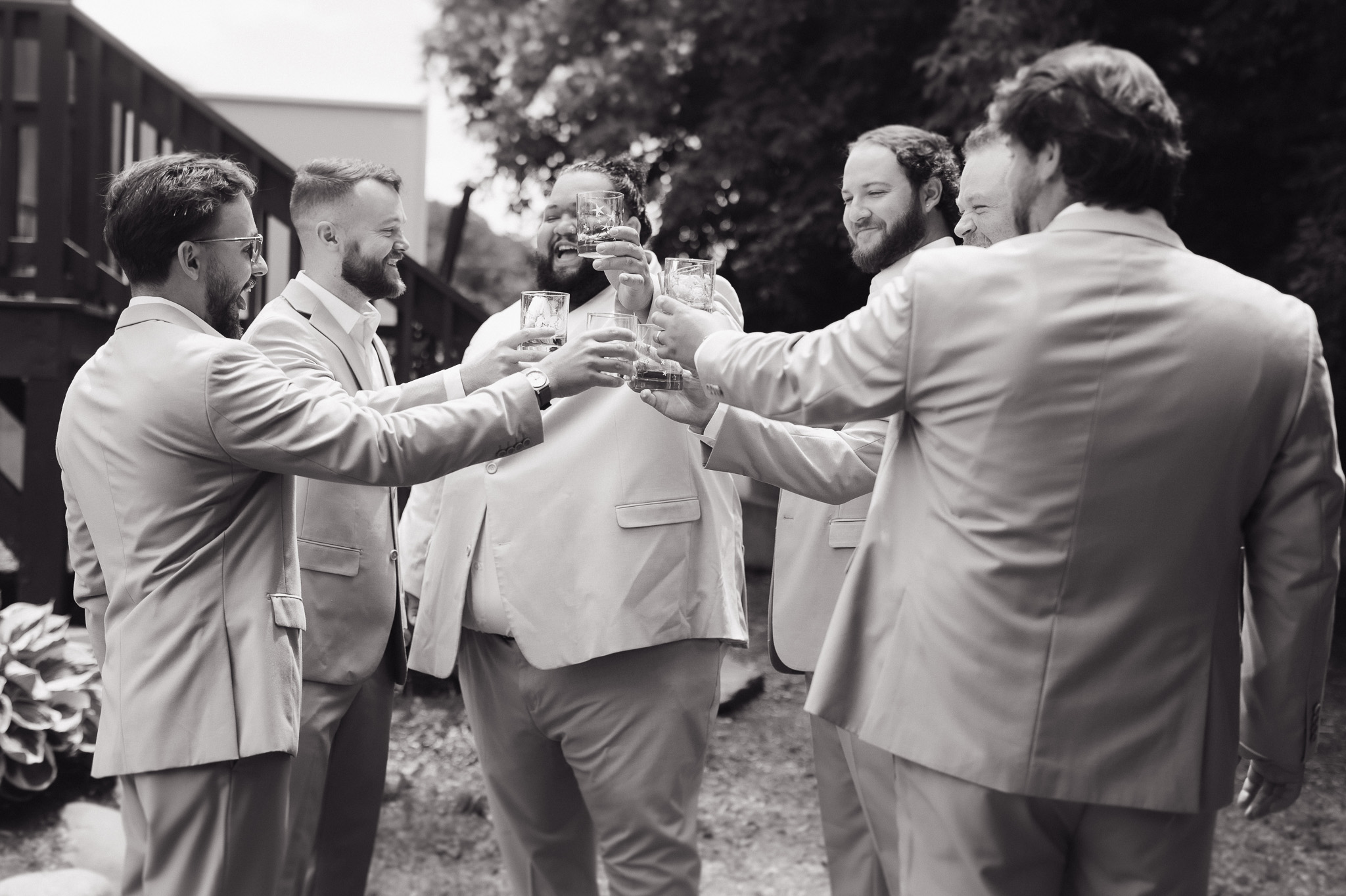 Groomsmen Wedding Day Toast Black-and-white candid photo of groomsmen in light neutral suits raising glasses together for a wedding day toast.